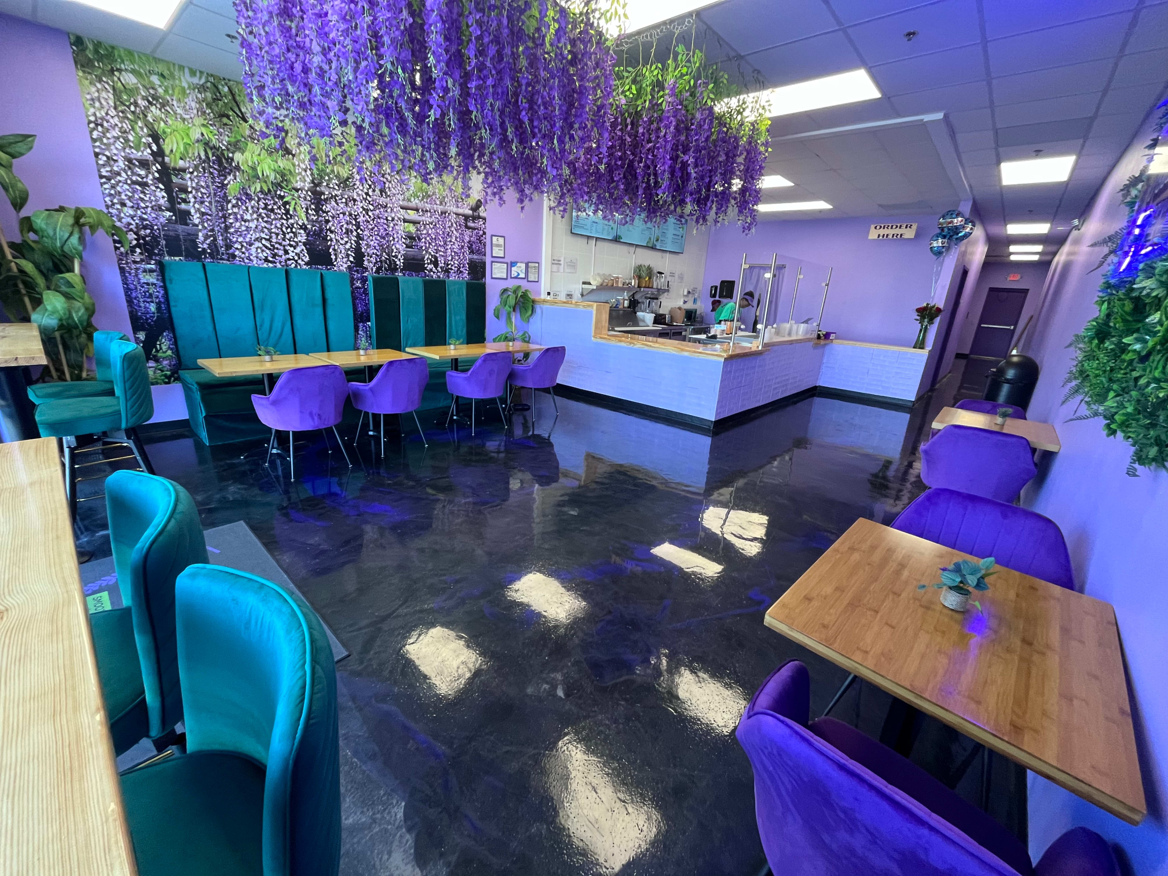 The image shows a brightly lit café with lavender walls, purple and teal seating, wood tables, and decorative plants, featuring a counter at the back.