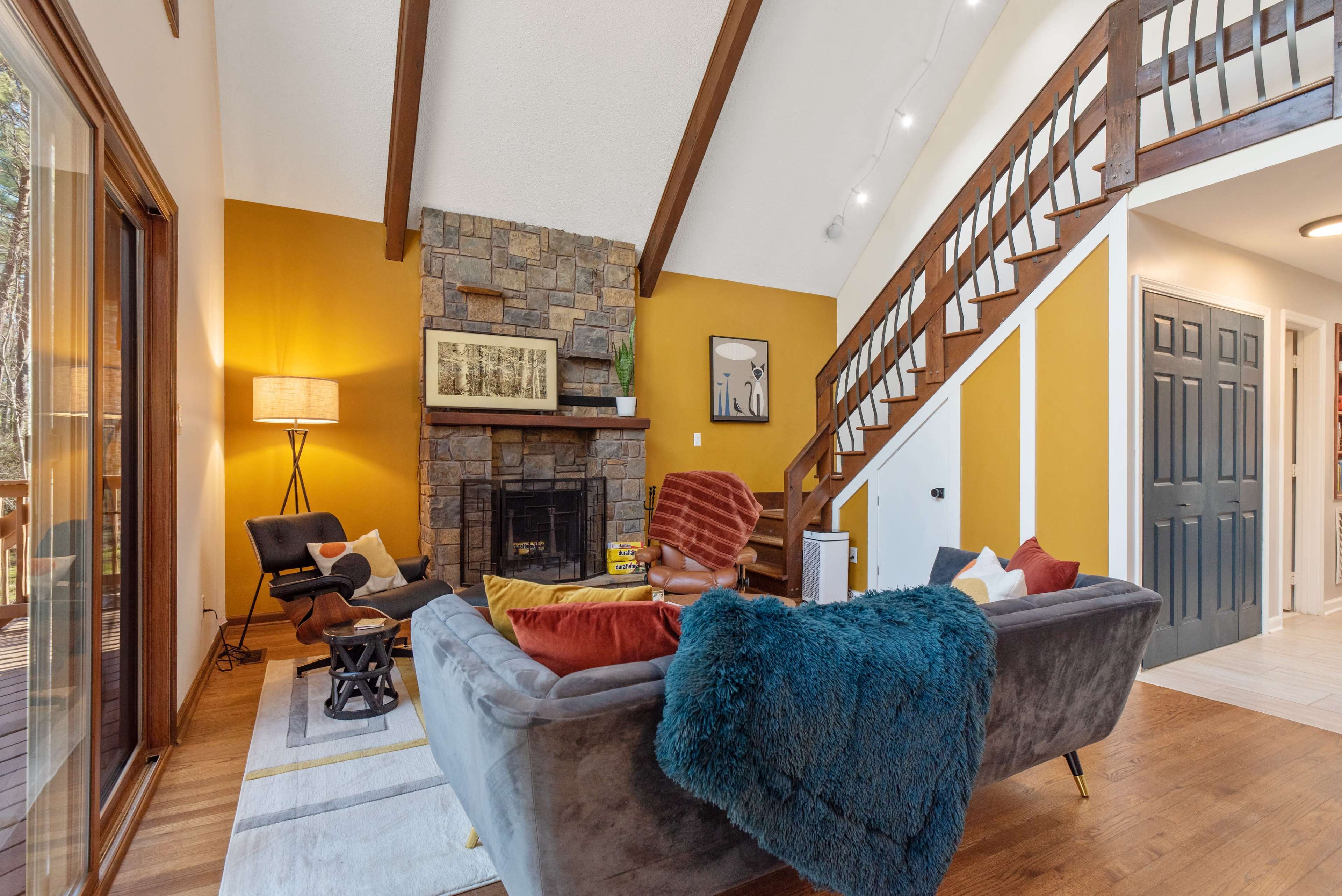 A cozy living room featuring a stone fireplace, a staircase with wooden railings, and a mix of modern and classic furniture.