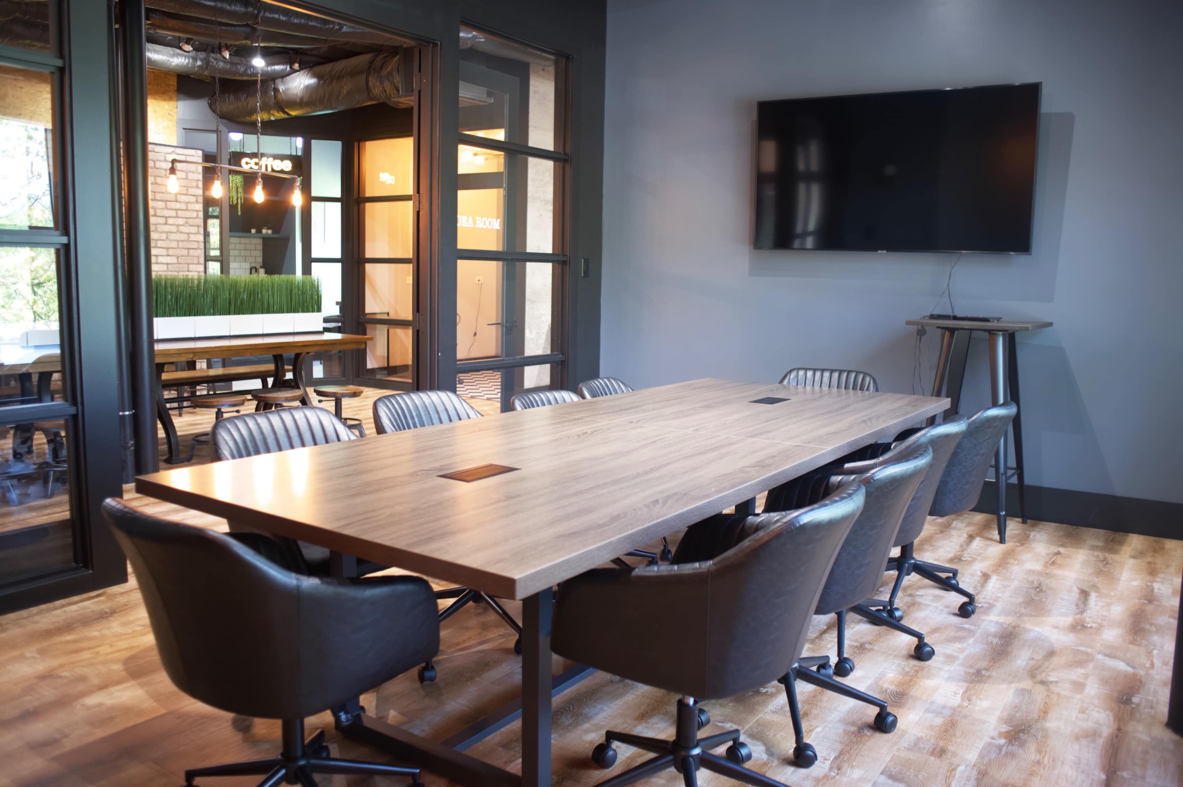 The image shows a modern conference room featuring a large wooden table surrounded by black chairs, with a wall-mounted screen and glass doors leading to another area.