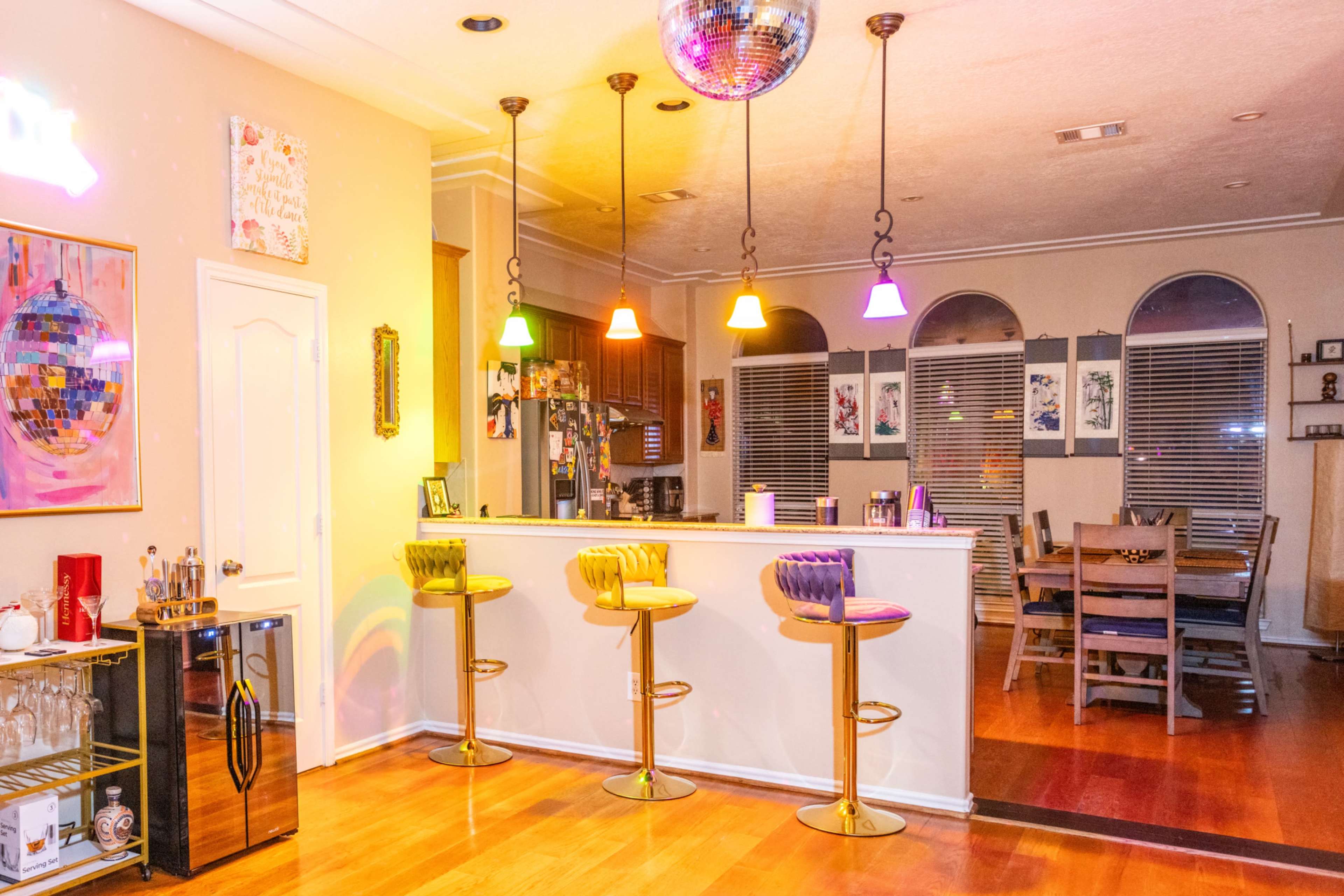 A modern kitchen and dining area features a bar with colorful pendant lights, high stools, and a dining table in the background.