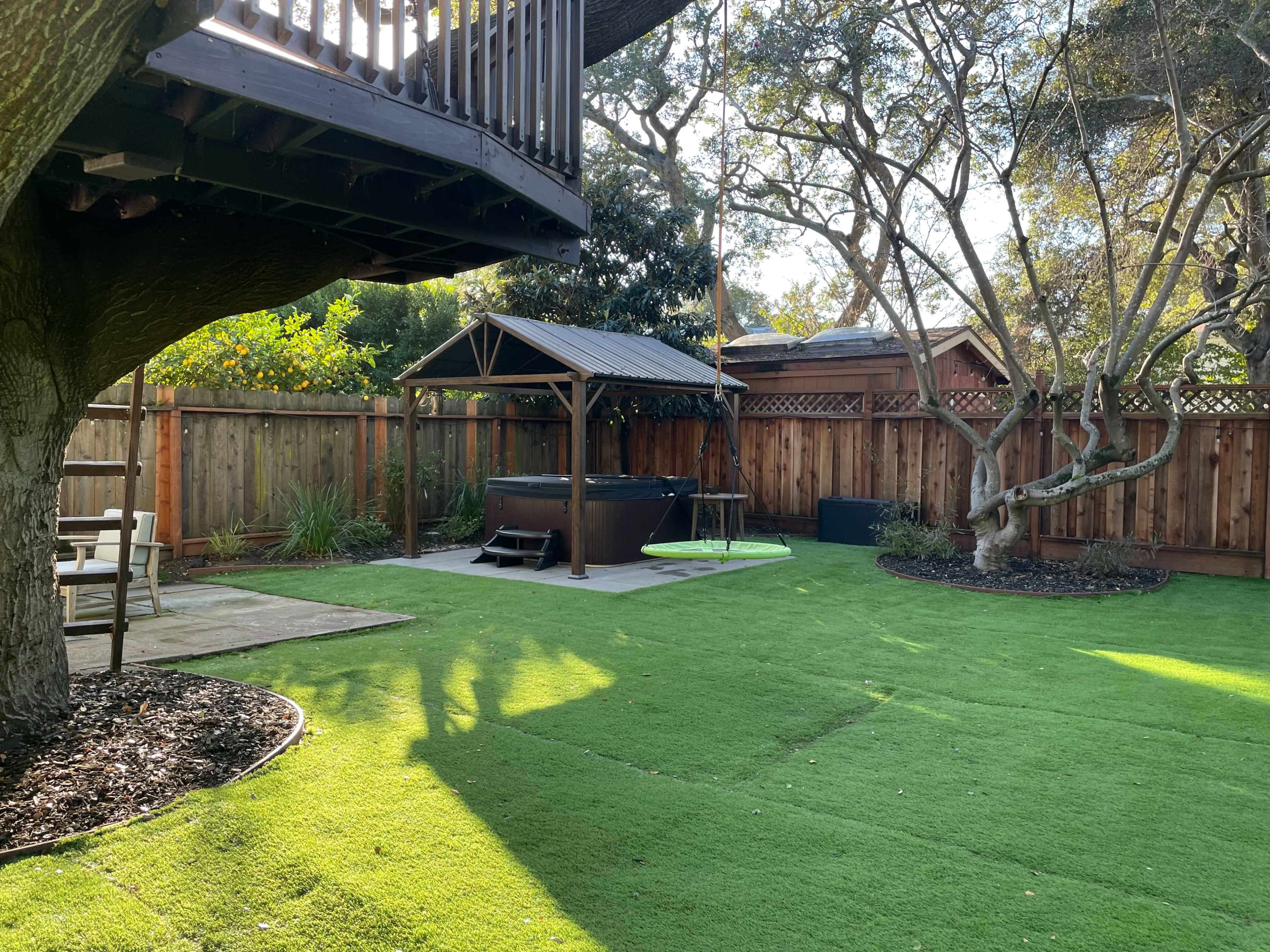 The image shows a backyard with artificial grass, a hot tub under a gazebo, and a wooden deck overhead surrounded by trees.