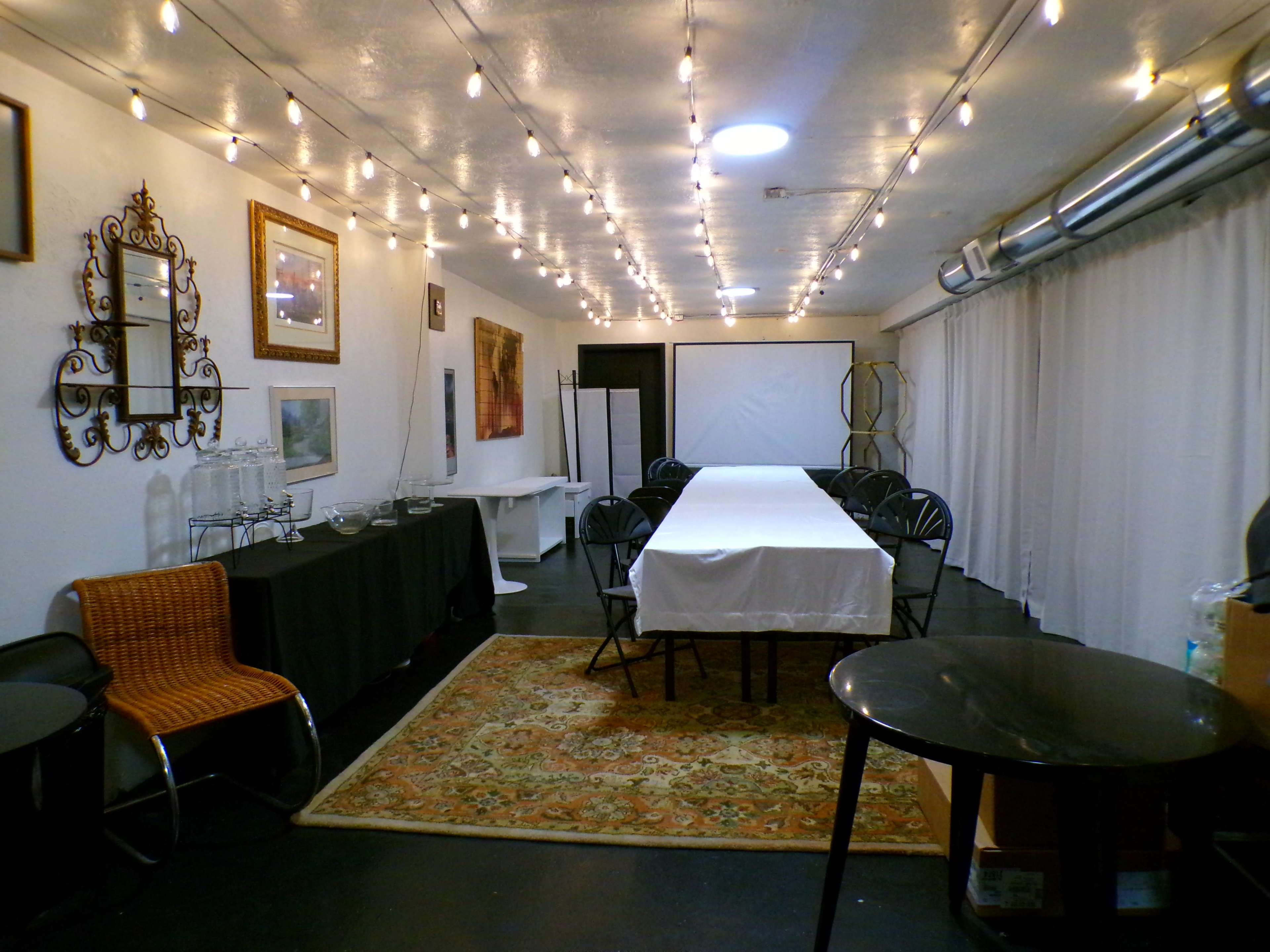The image shows a rectangular room decorated with string lights, featuring a long table in the center, surrounded by black chairs, with a carpet on the floor and various furnishings along the walls.
