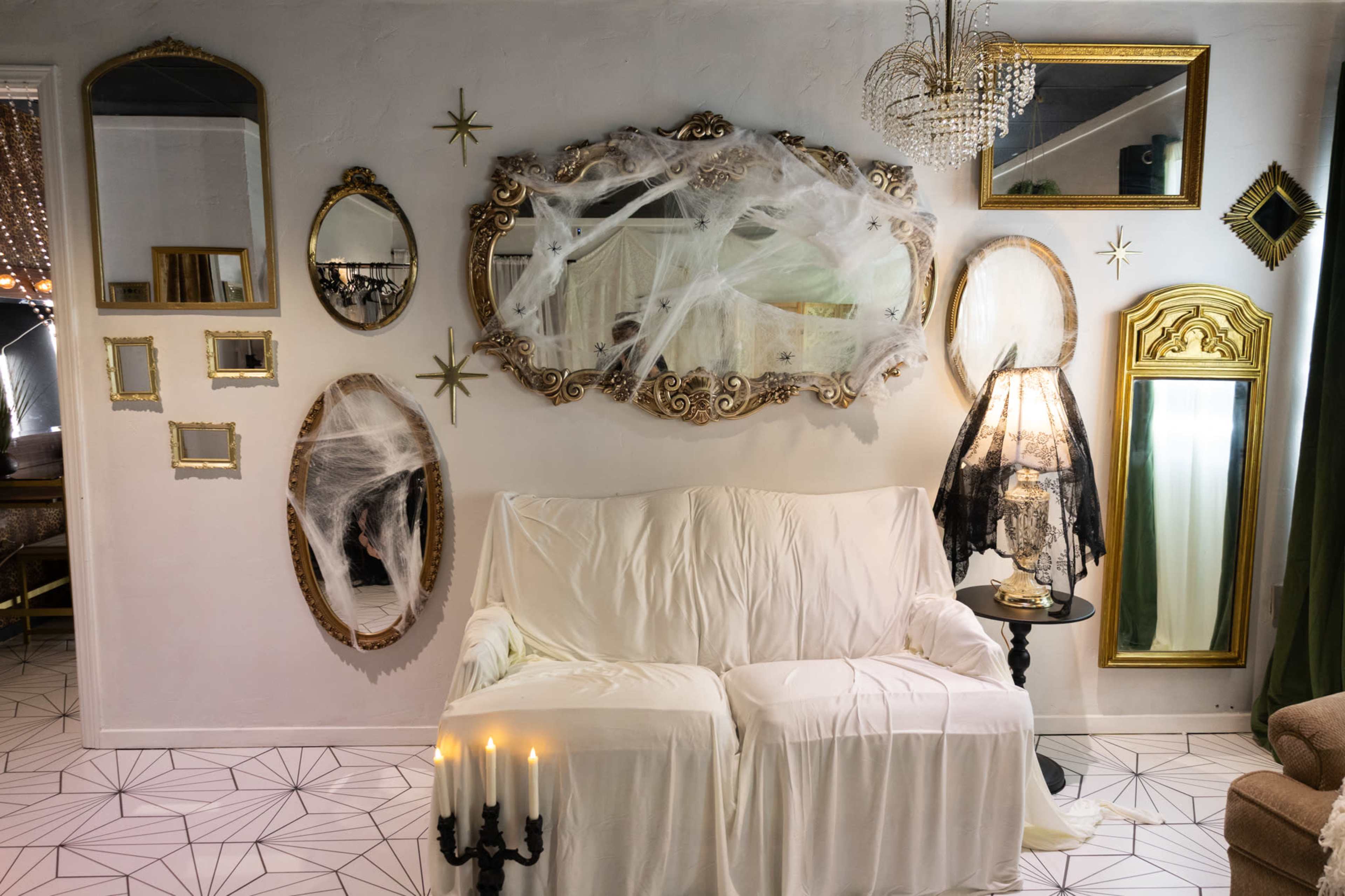 The scene features a sofa covered with a white cloth in front of a wall adorned with various ornate mirrors and spider web decorations, along with a lamp and candles.