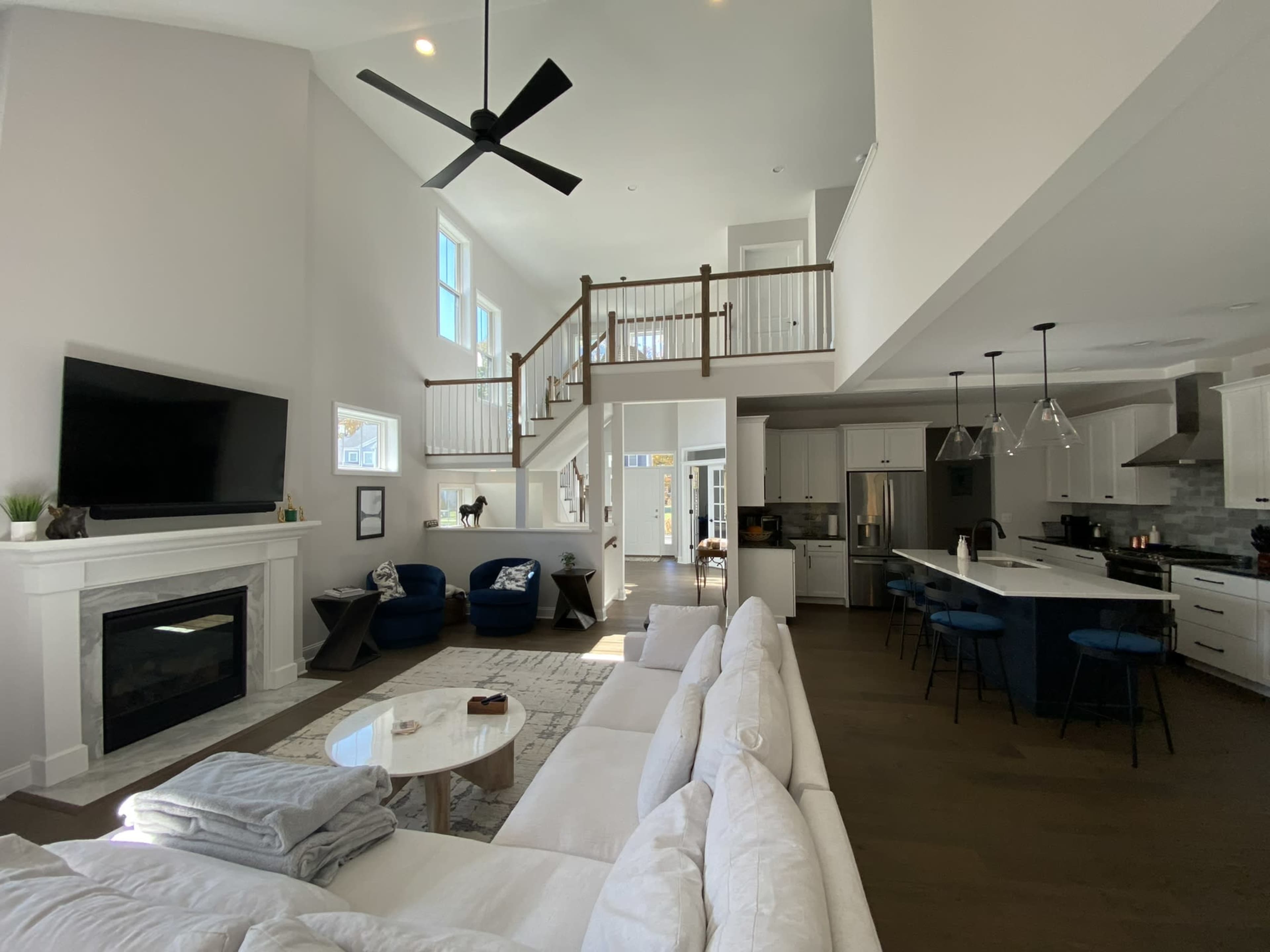 A spacious and modern living area with a white sectional sofa, a large flat-screen TV mounted above a fireplace, and an open kitchen featuring an island with bar seating.