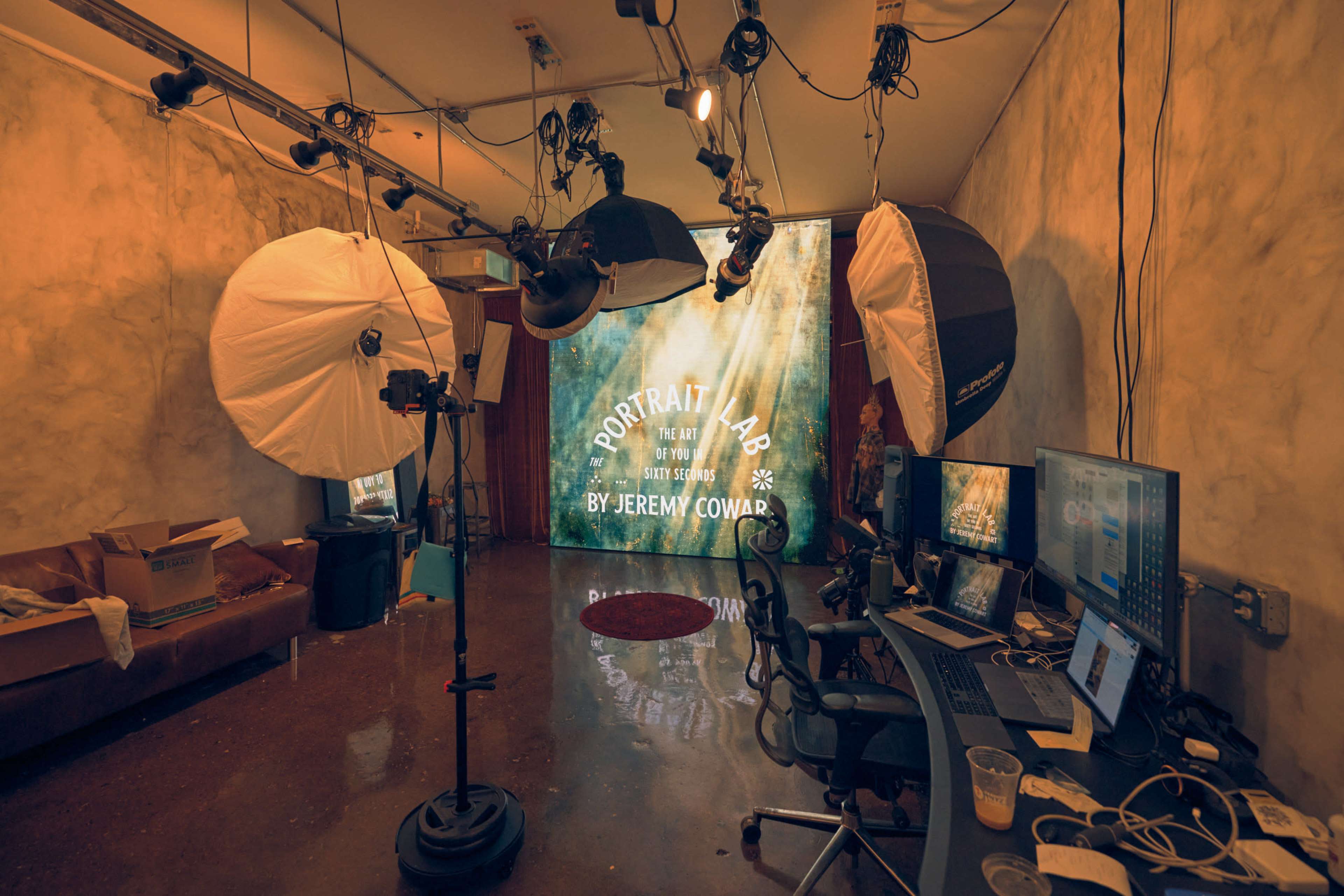 The image shows a photography studio equipped with multiple light umbrellas, camera tripods, and a computer workstation against a textured wall.