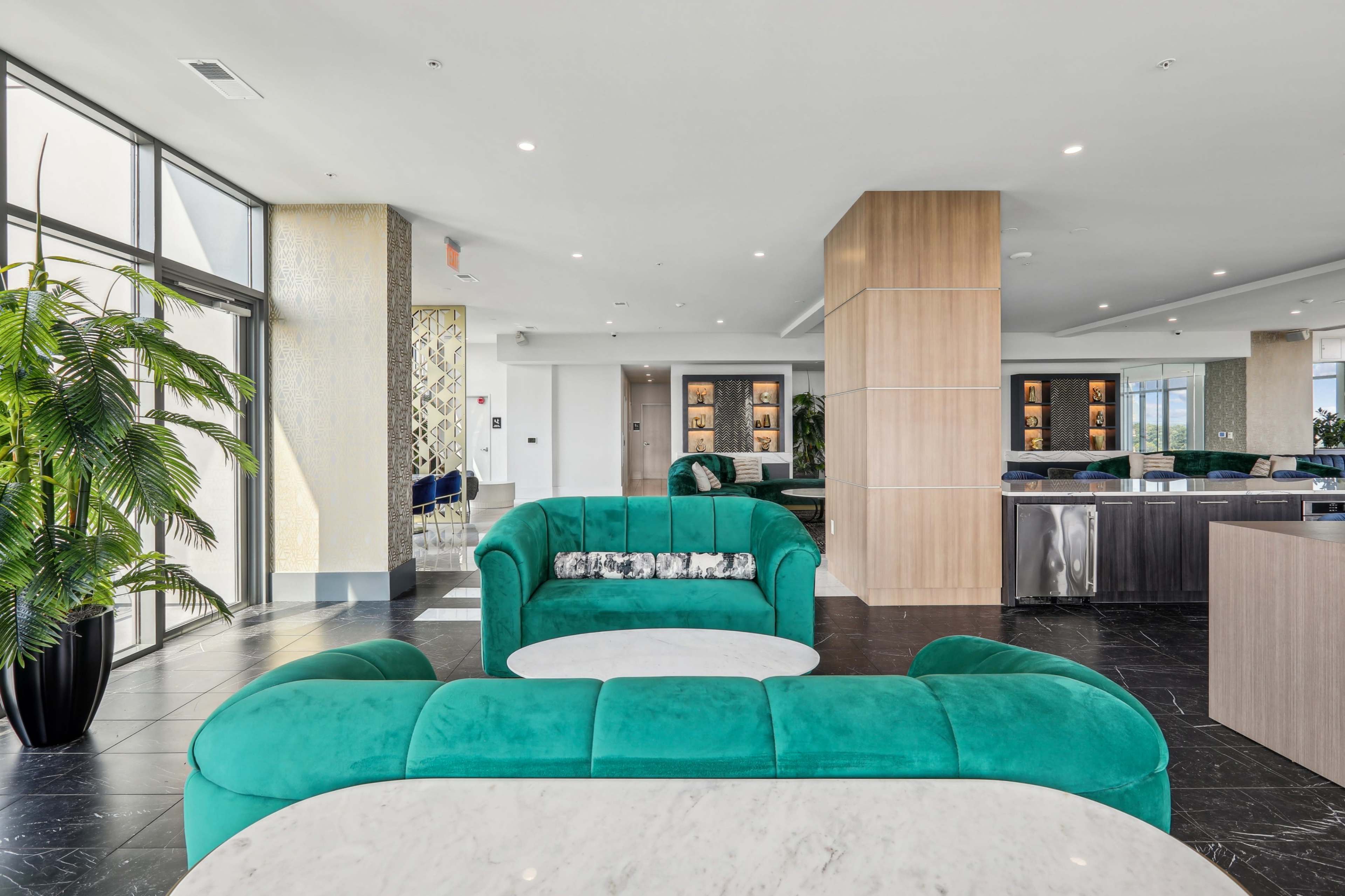 A modern lounge area features aqua-colored sofas and marble tables, with large windows and lush greenery.