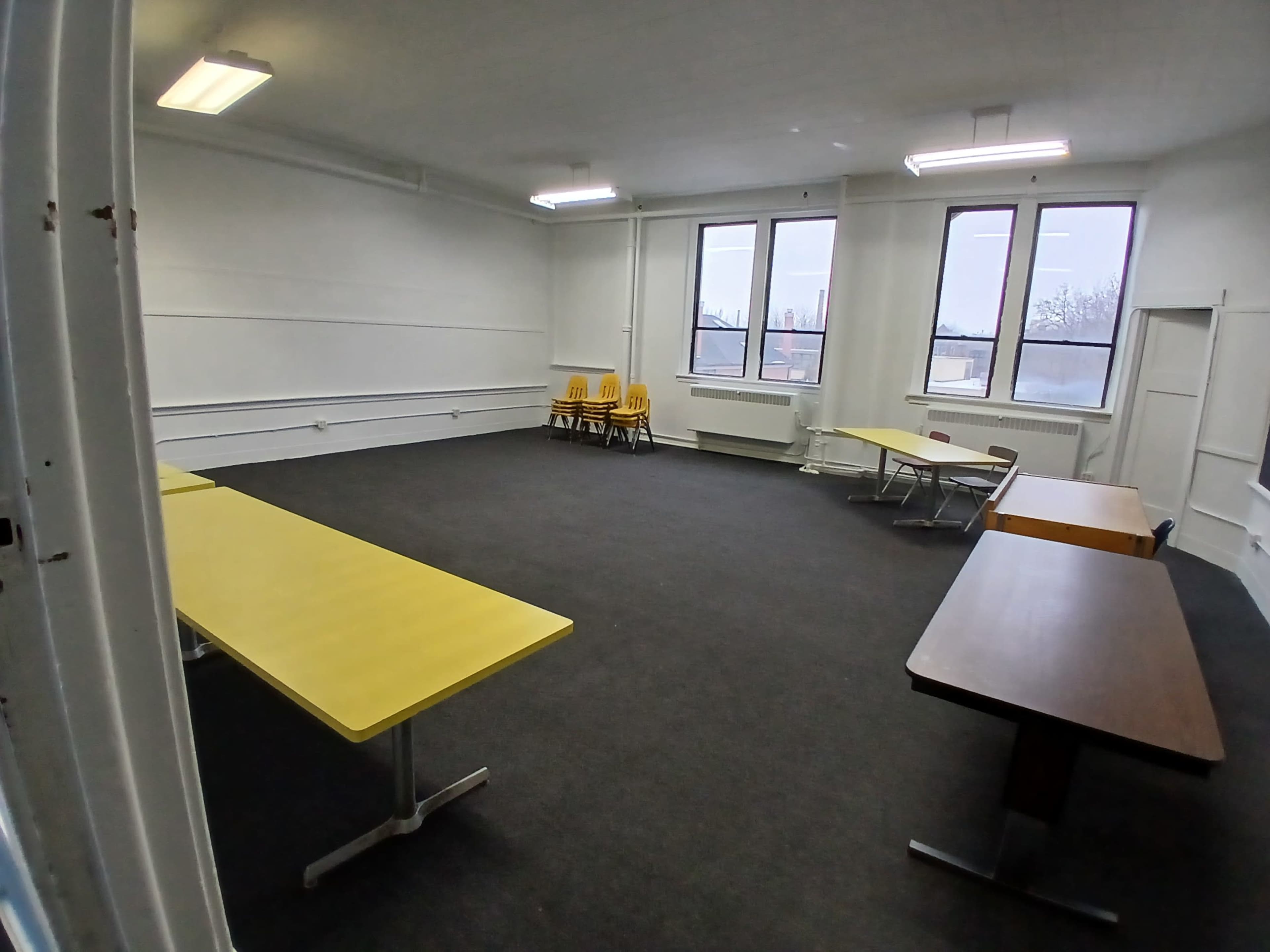 A sparsely furnished classroom features several tables and a stack of yellow chairs under bright overhead lights.