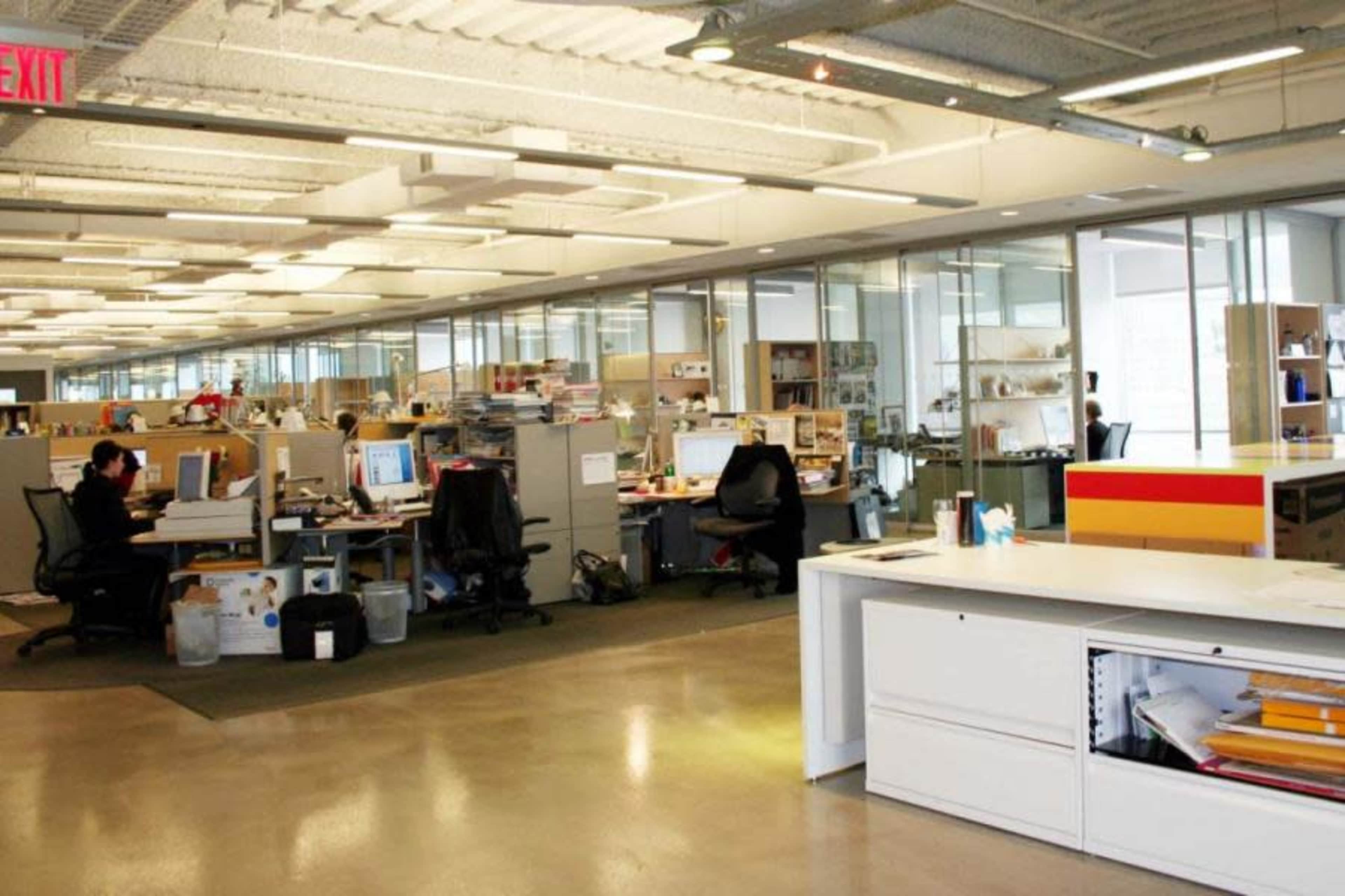 An open office space with cubicles, desks, computers, and large windows allowing natural light.