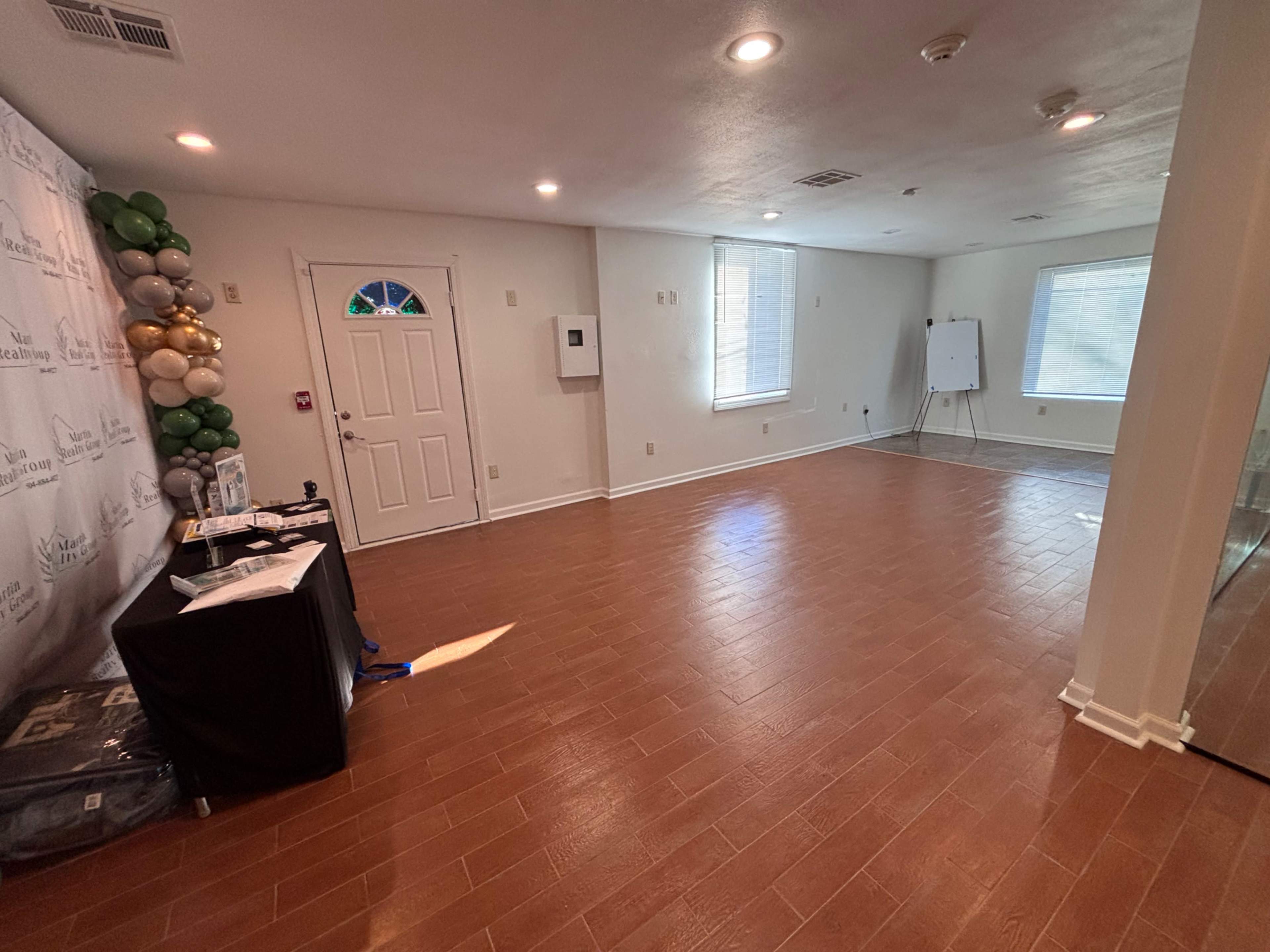 The image shows an empty room with hardwood flooring, a door, two windows, and a small table set up against one wall.
