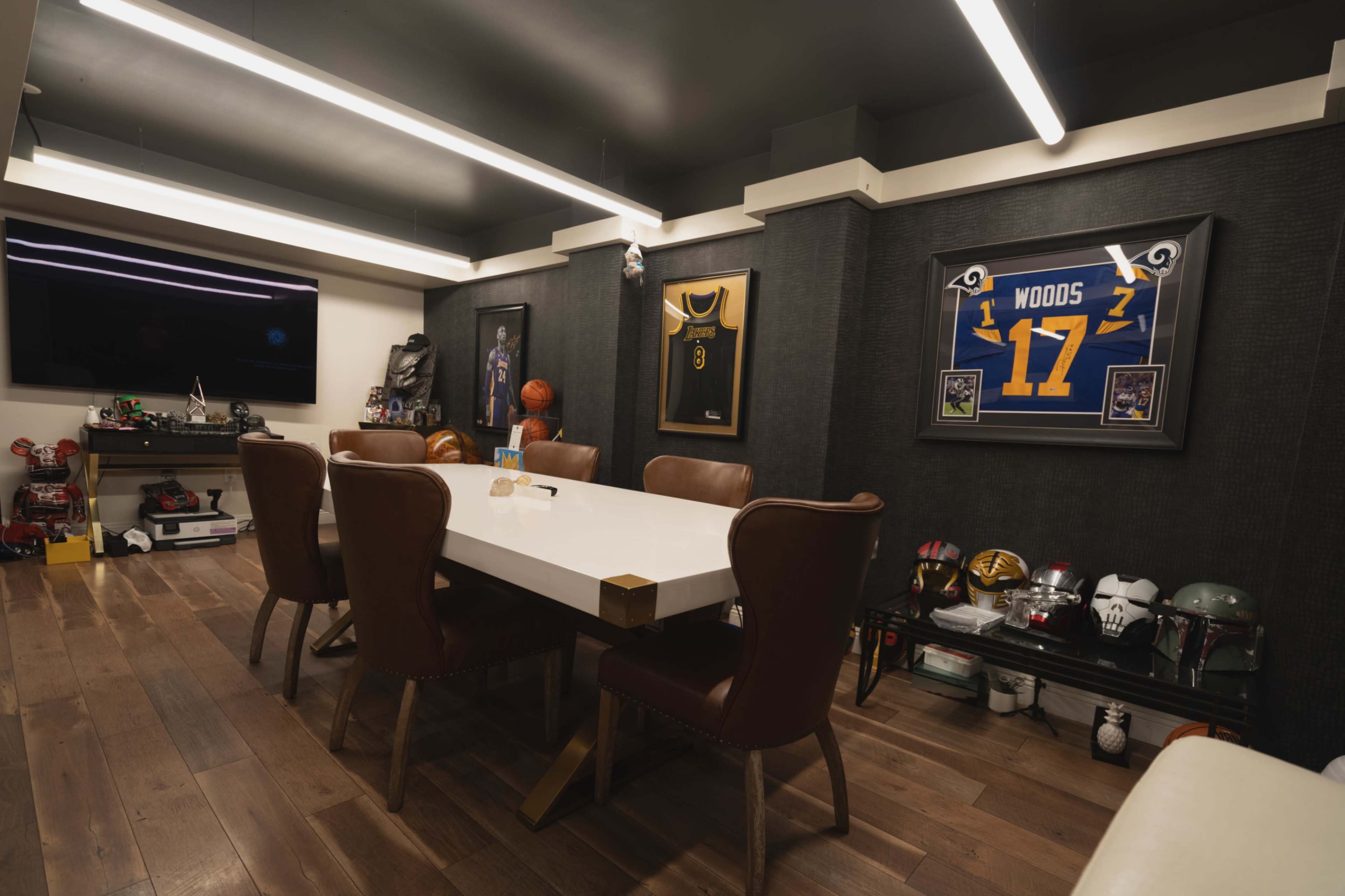 The image shows a modern dining area with a white table and brown upholstered chairs, framed sports jerseys on the walls, and various collectibles displayed on shelves.