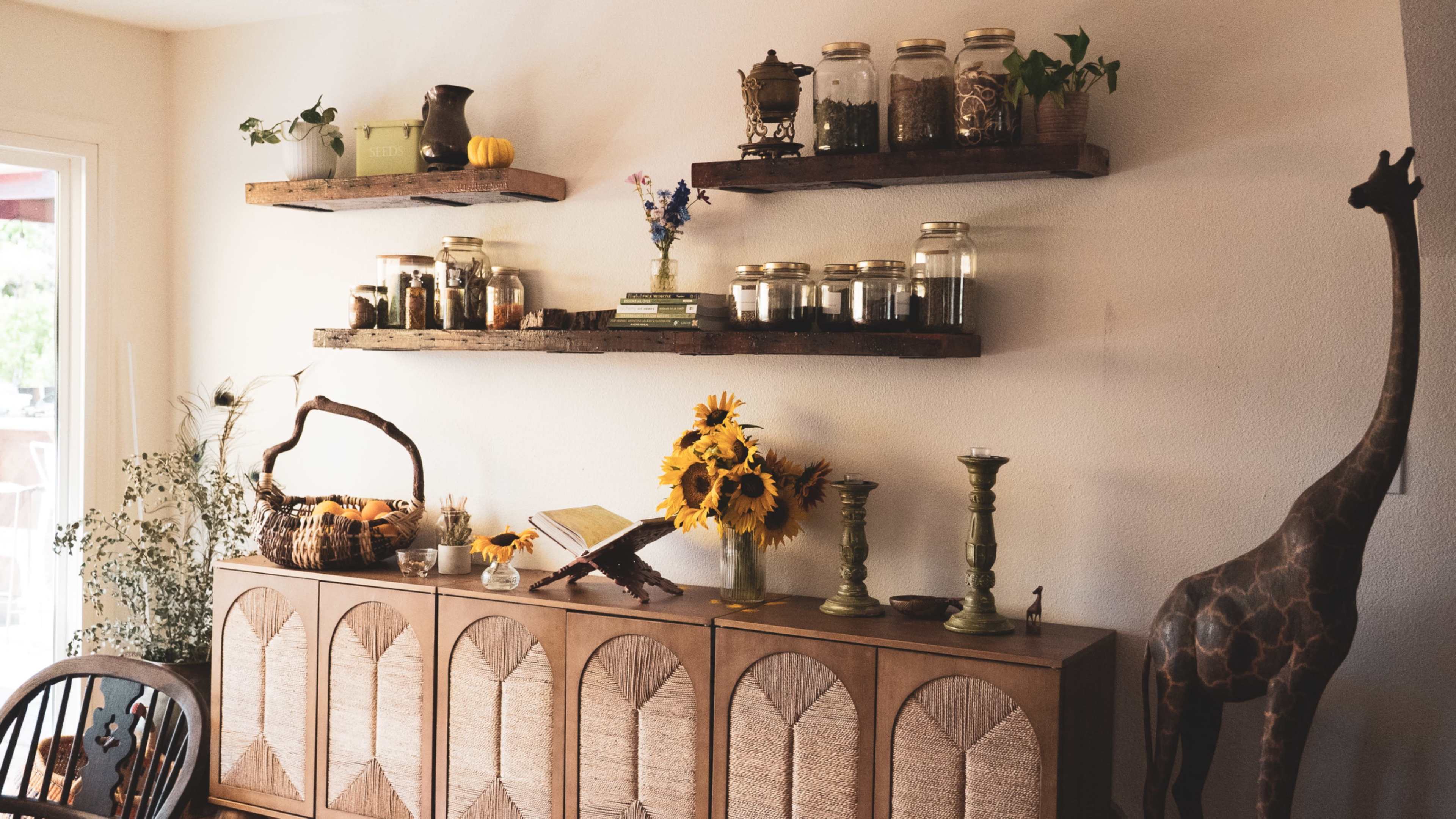The scene features a wall with wooden shelves displaying jars, plants, and decorative items above a cabinet adorned with a basket, sunflowers, and candlesticks, alongside a giraffe figurine.