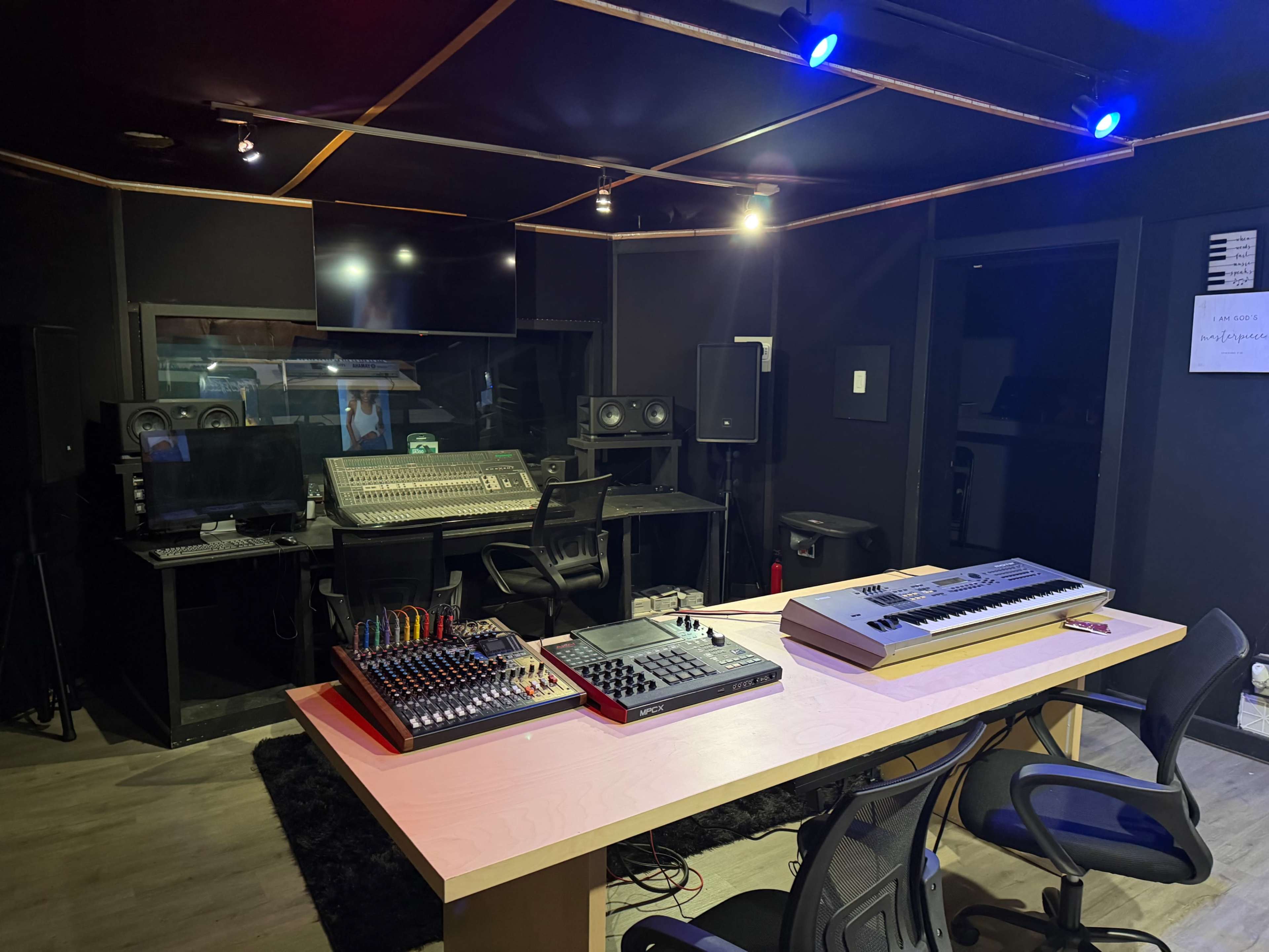 The image shows a modern music studio featuring a mixing console, audio equipment, and a desk with a keyboard and music production devices.