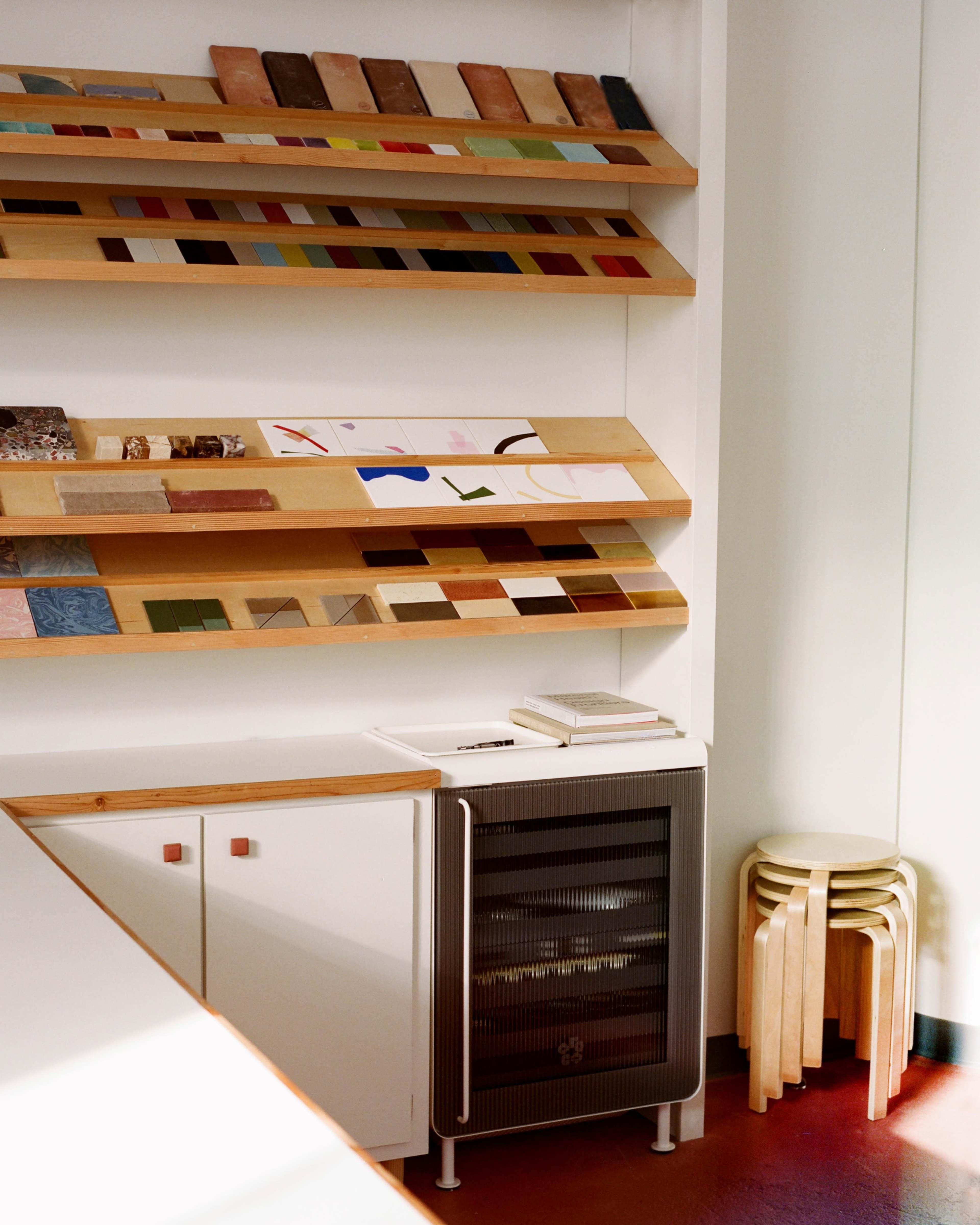 The image shows a design space with a wall-mounted display of color samples and materials, alongside a small fridge and a set of wooden stools.