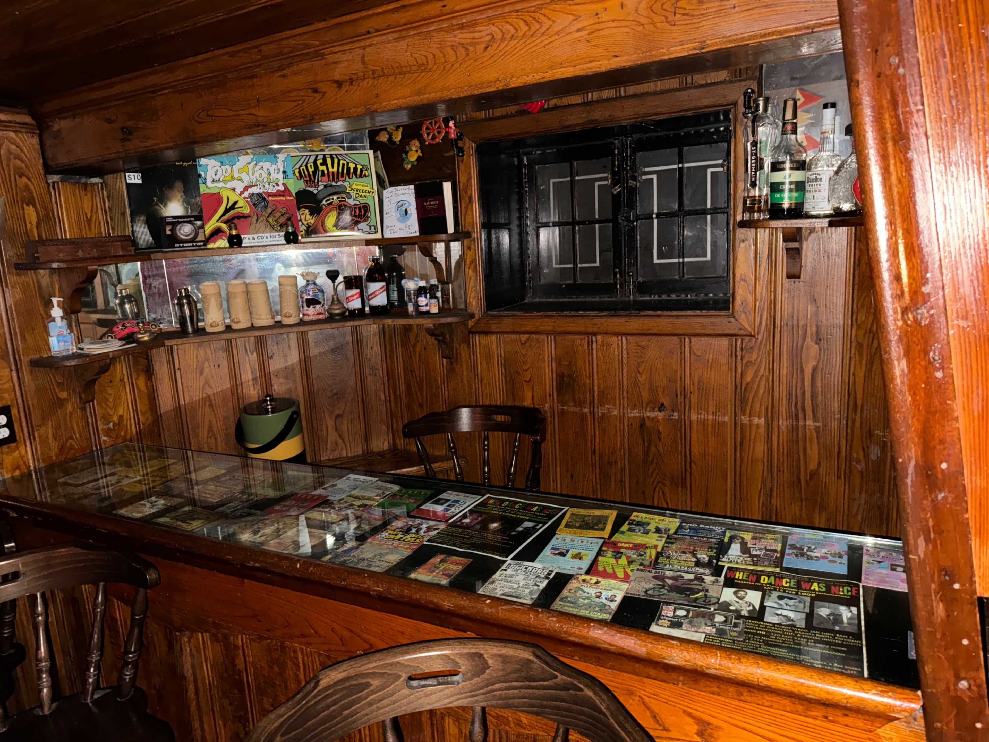 A wooden bar with a glass top displaying various flyers and magazines, surrounded by shelves holding bottles and a large window.