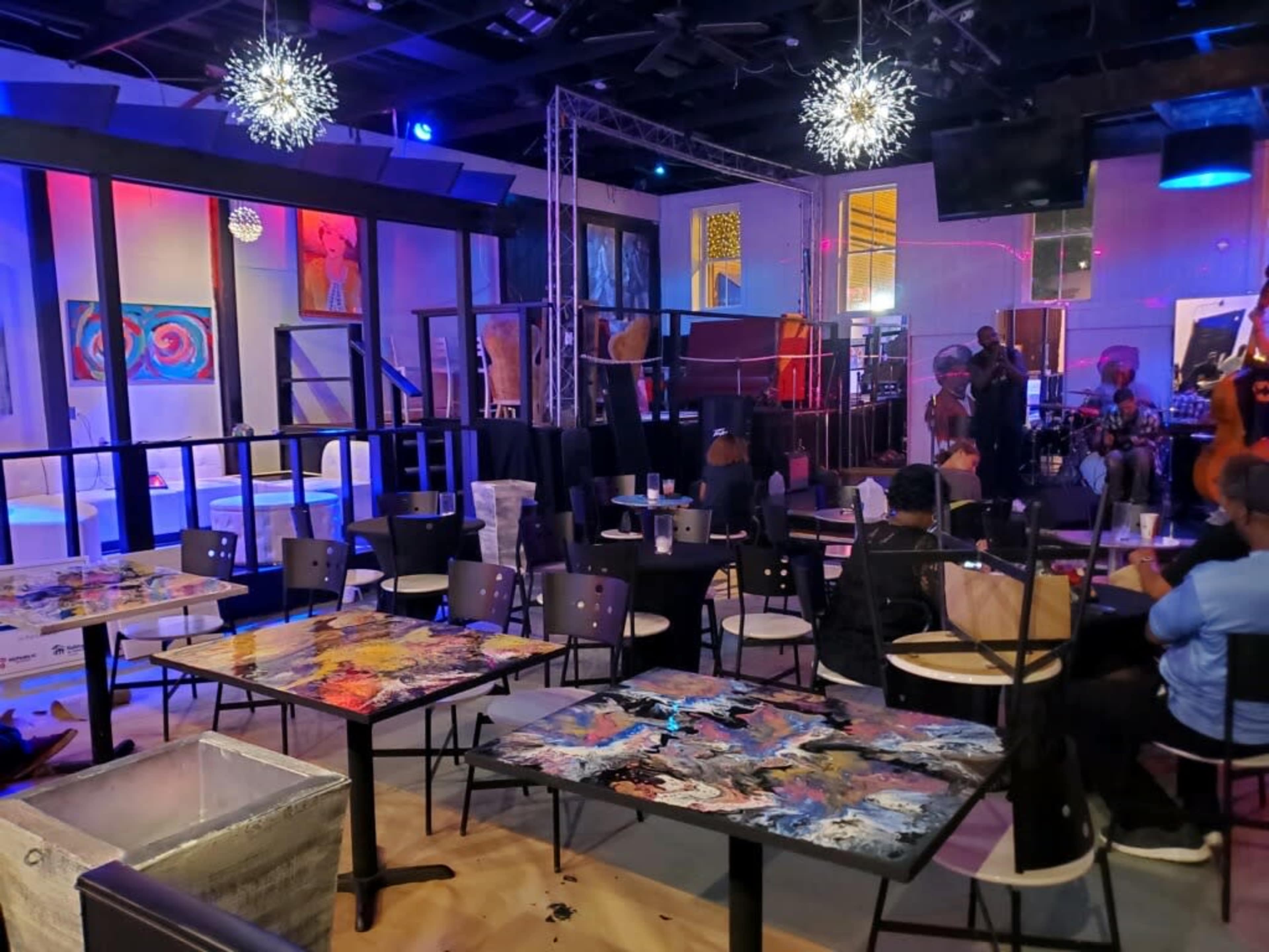 A lively indoor venue with colorful artwork displayed, tables covered in paint, and a small stage where a band is performing.