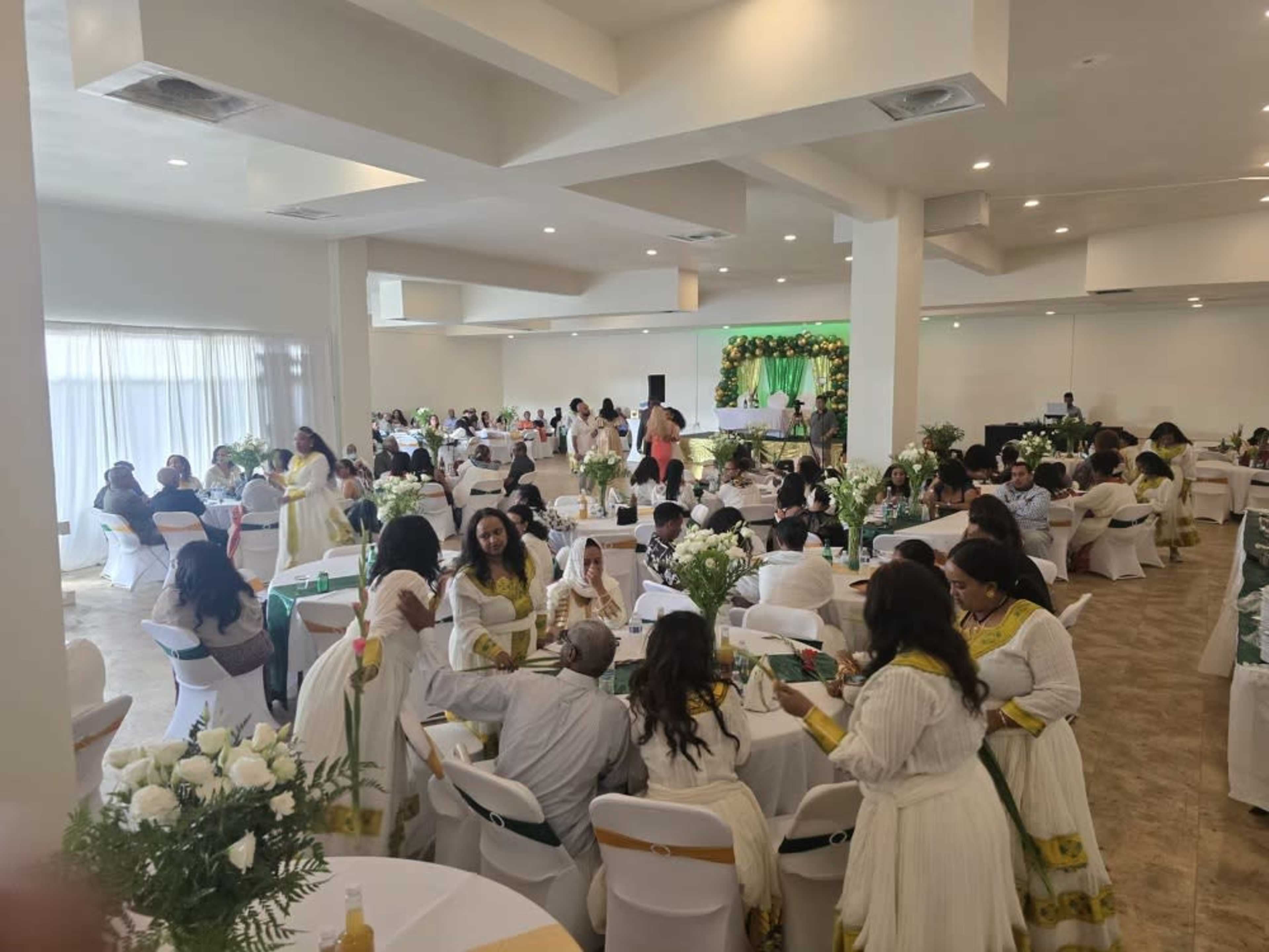 The image depicts a banquet hall filled with guests seated at tables, dressed in white attire, gathered for a celebration with floral arrangements and decorations in the background.