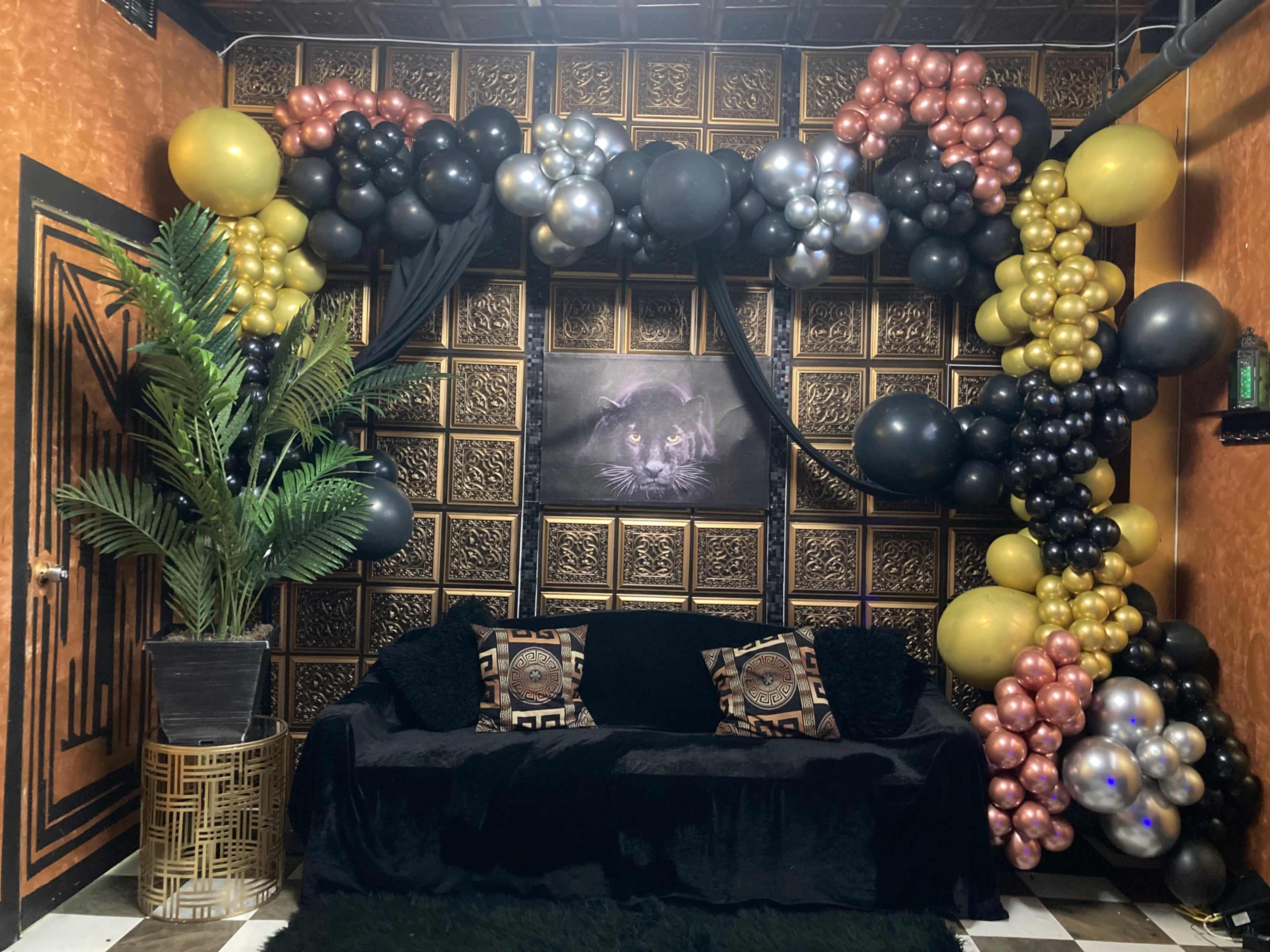 The image shows a decorated corner of a room featuring a black couch with decorative pillows, a large plant, and an archway of balloons in black, gold, and silver against a textured wall.