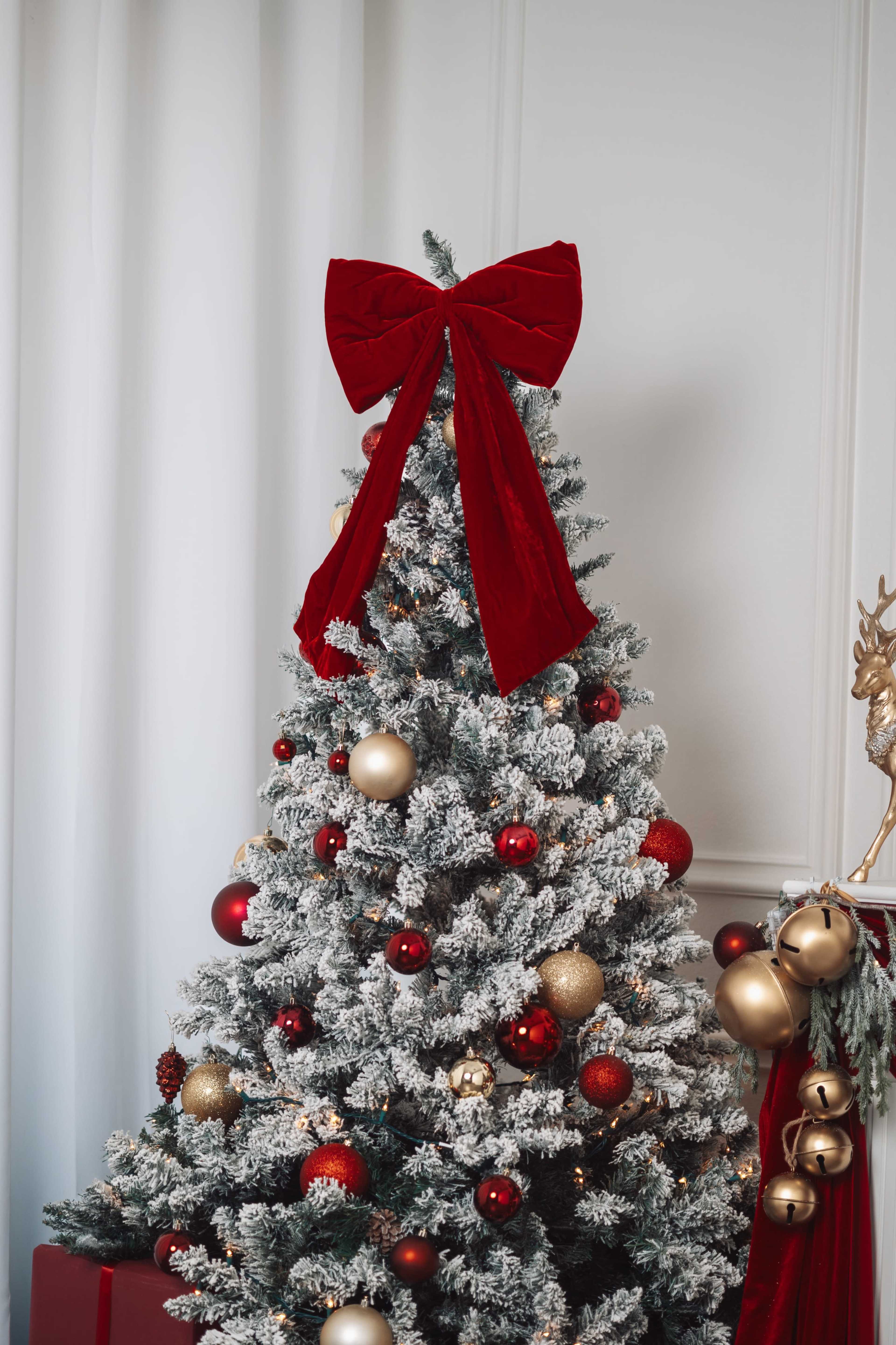 A decorated Christmas tree with a large red bow on top stands in a room with white curtains.