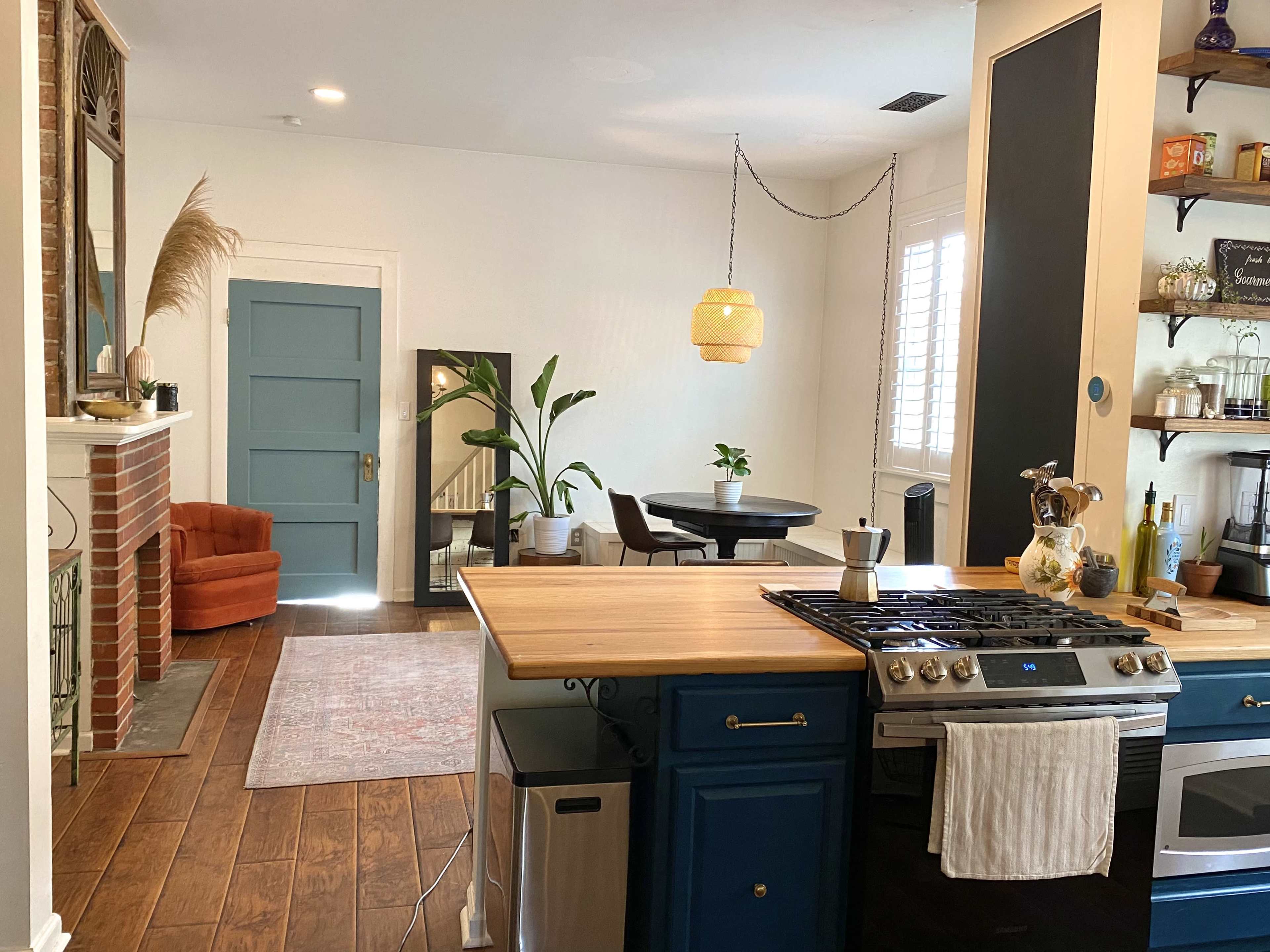 The image shows a modern kitchen with a wooden island, a gas stove, and a view into a cozy living area featuring a brick fireplace, an orange chair, and a round dining table.