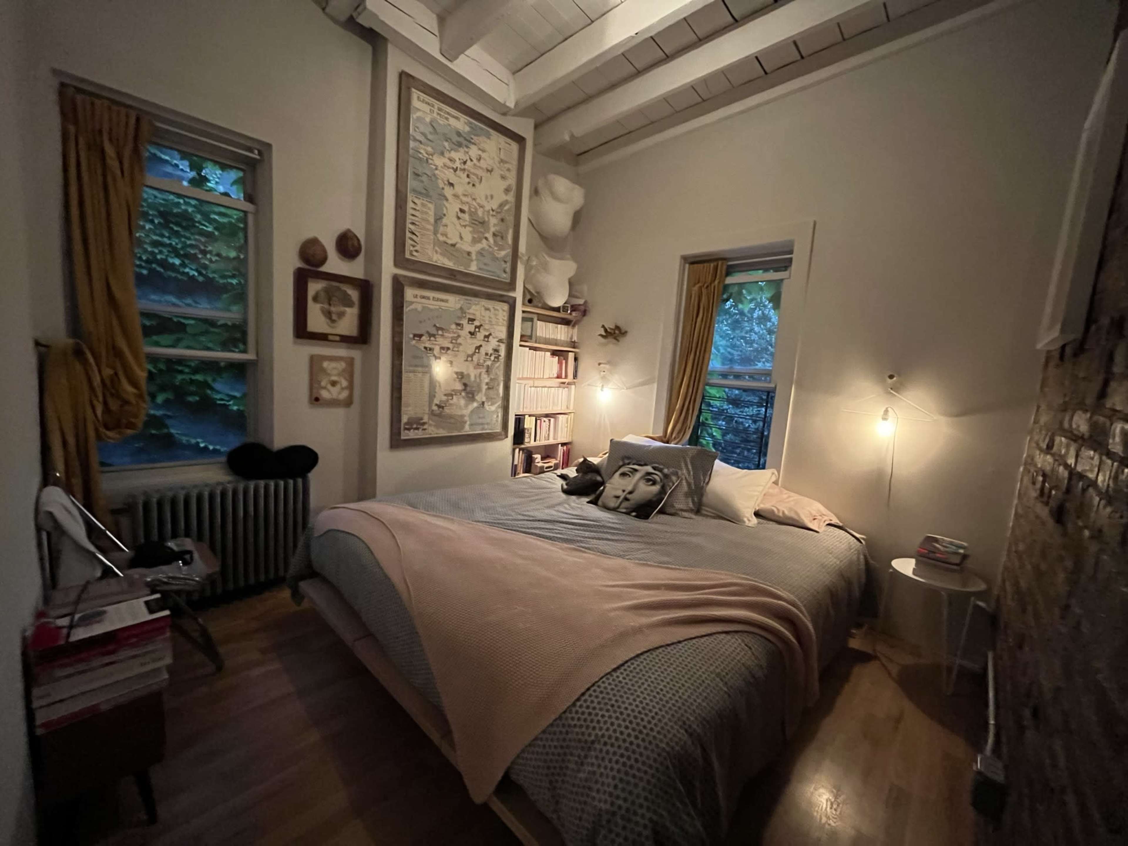 The image shows a cozy bedroom with a large bed, bookshelves, and maps hanging on the walls, illuminated by soft lighting from a bedside lamp and natural light from a window.