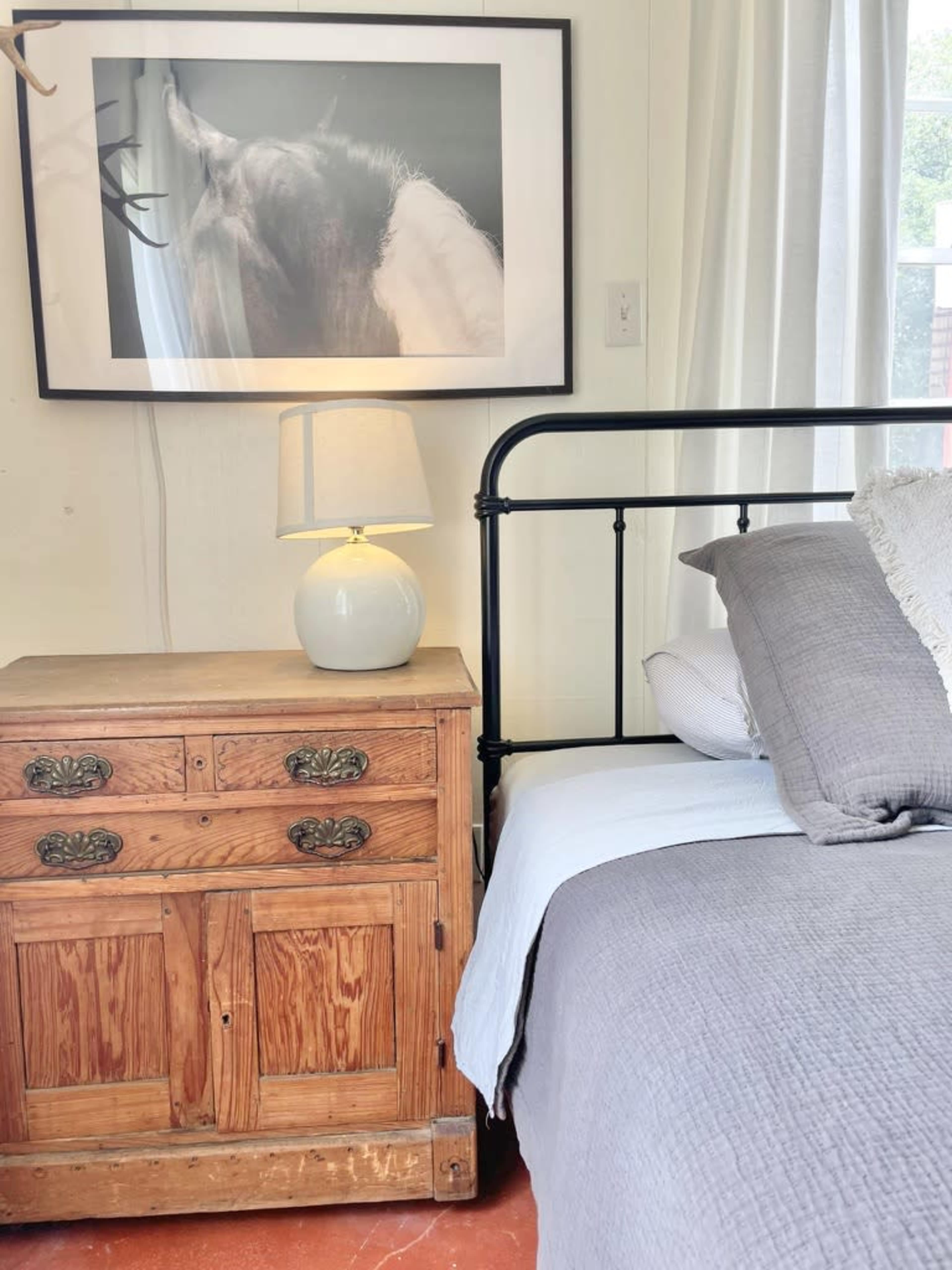 A bedside table with a white lamp next to a bed covered with gray bedding, framed by a large art piece of a horse on the wall behind it.