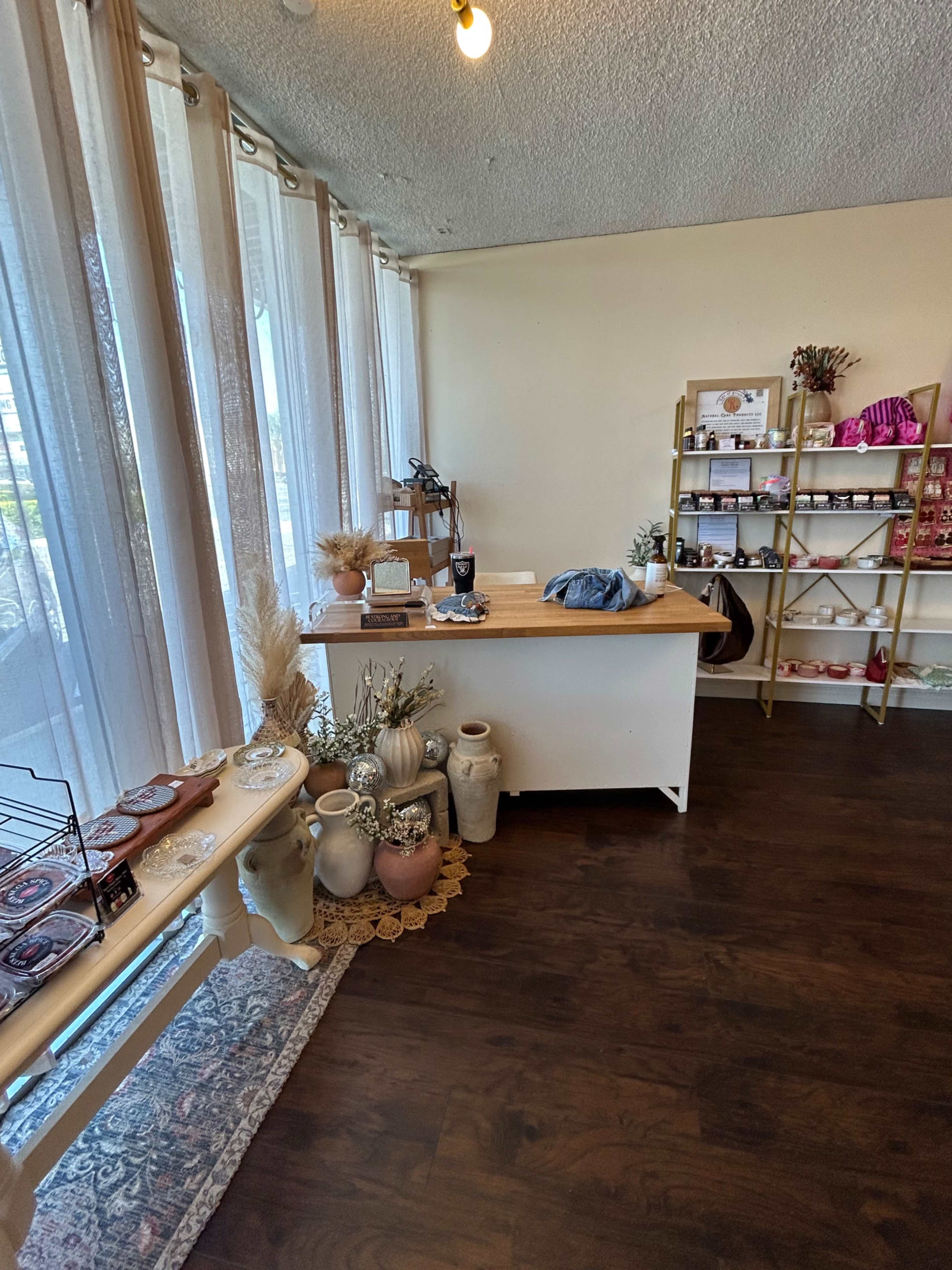 The image shows a cozy shop interior featuring a wooden counter with decorative pottery, a shelf displaying various products, and large windows allowing natural light to enter.