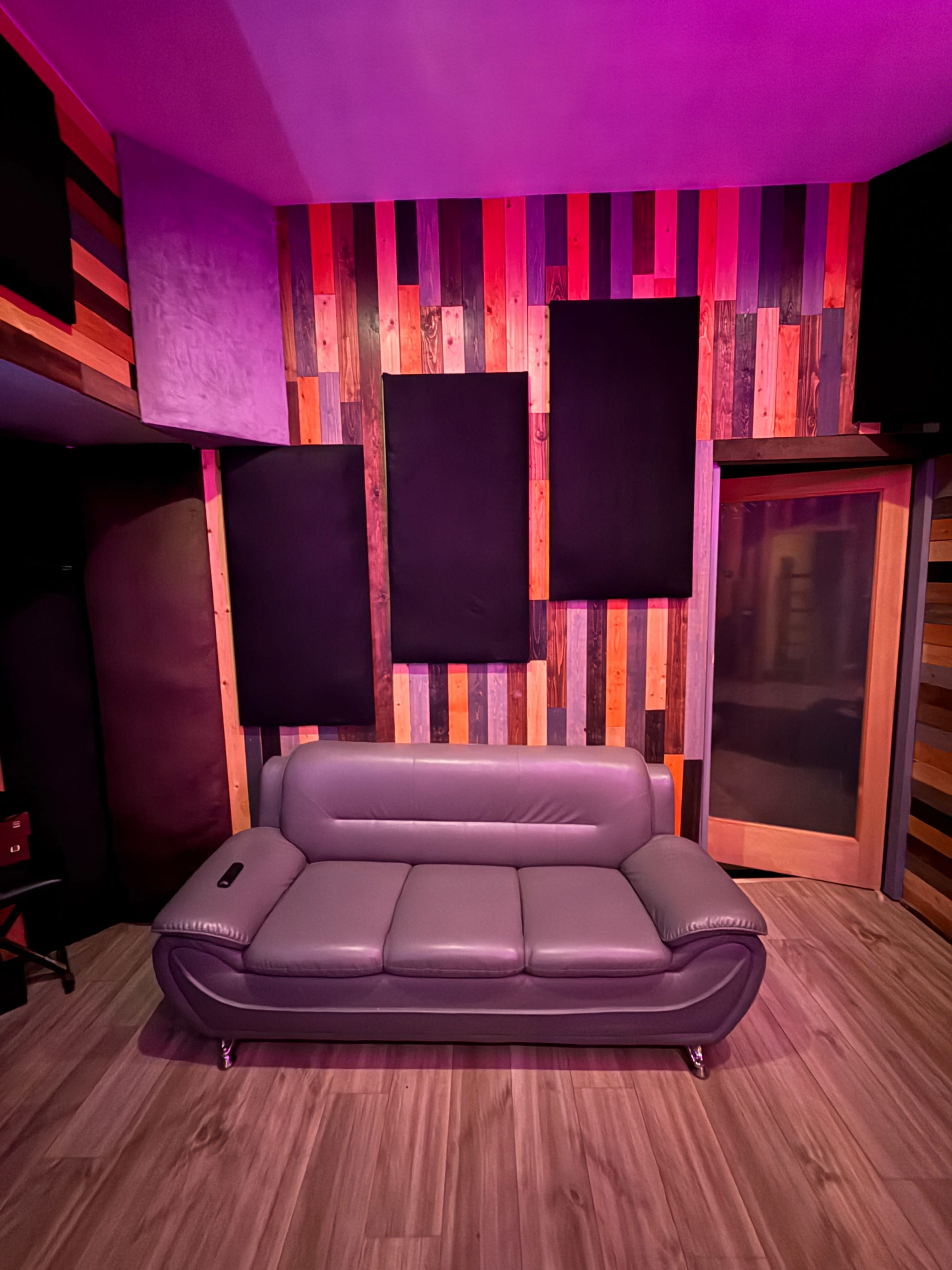 The image shows a modern lounge area with a gray leather sofa against a wall featuring colorful wooden panels and soundproofing elements.