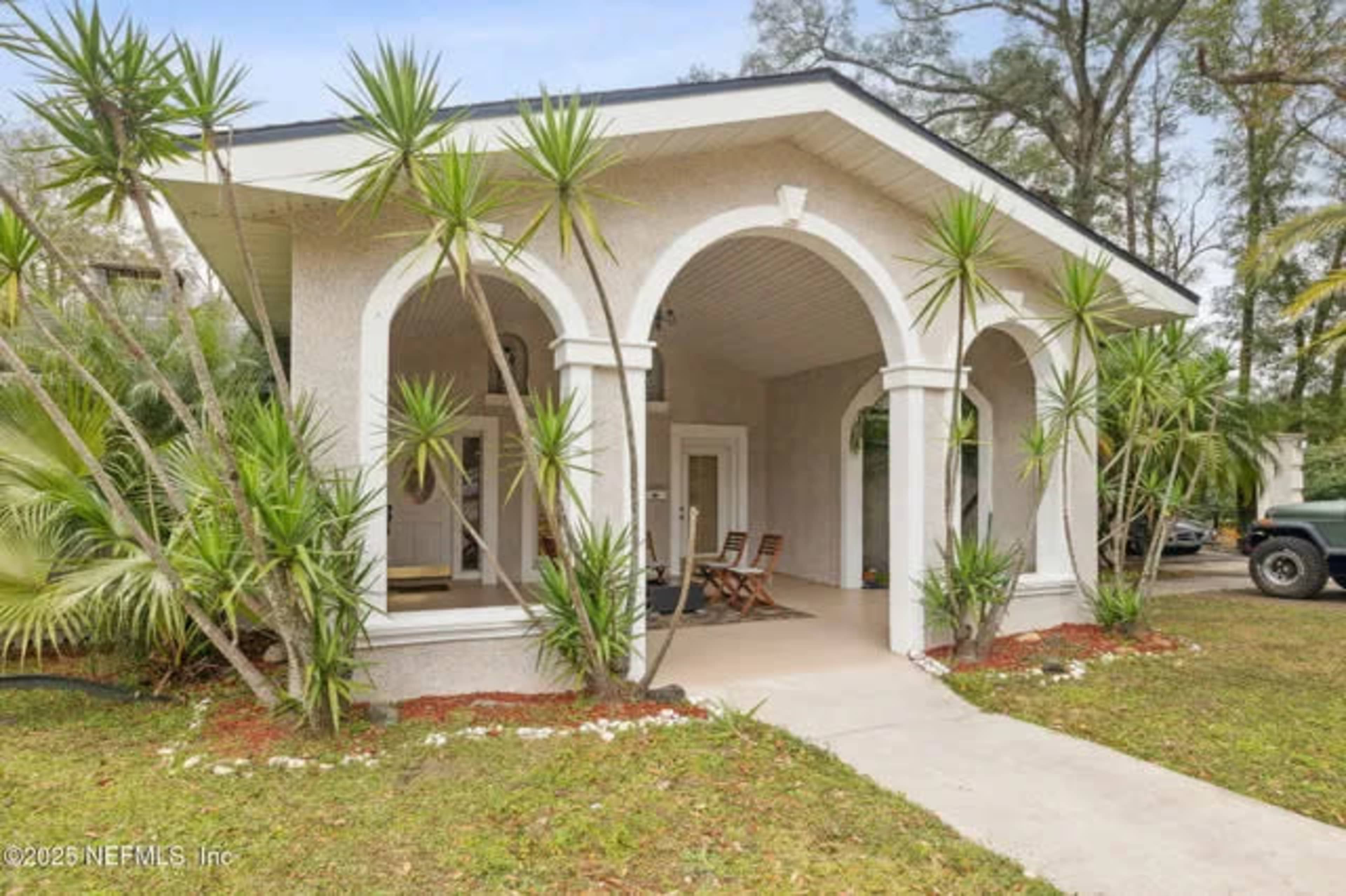 The image shows a house with a covered porch framed by arched columns and surrounded by palm plants.
