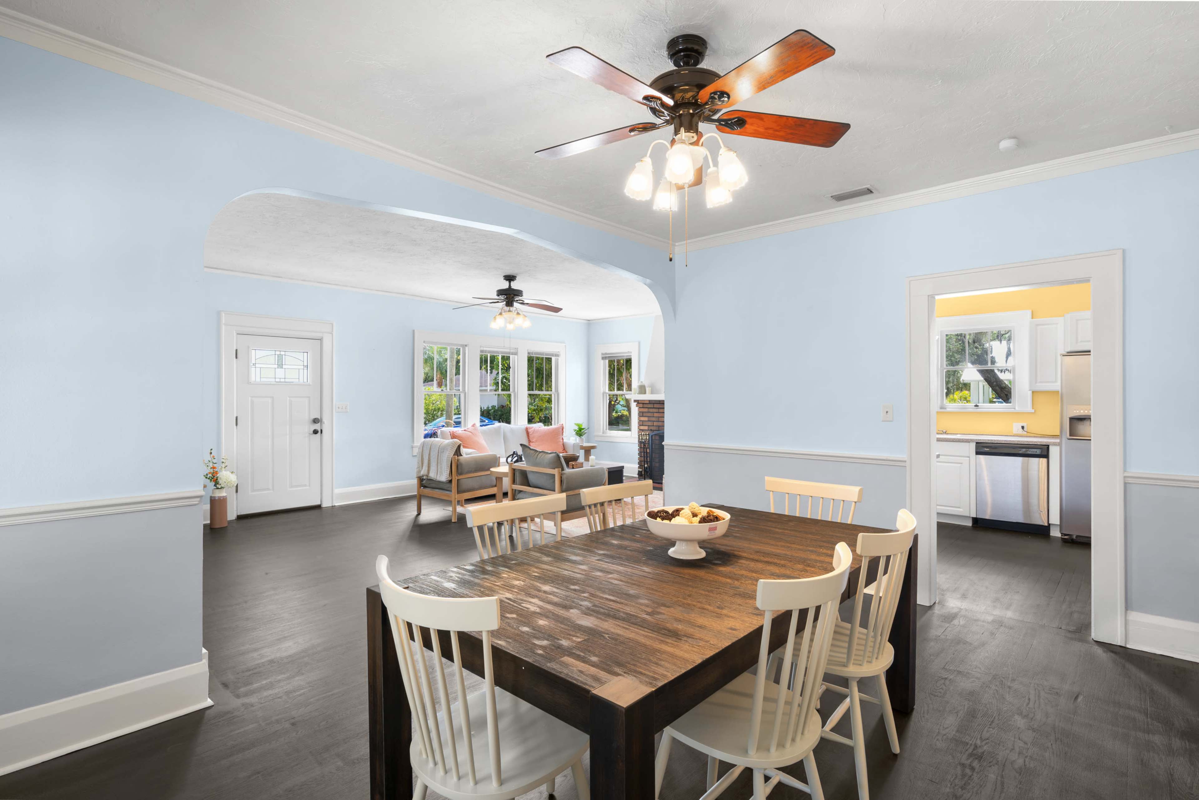 A spacious, light-colored dining area features a wooden table surrounded by white chairs, with a view of a living area and a kitchen beyond.