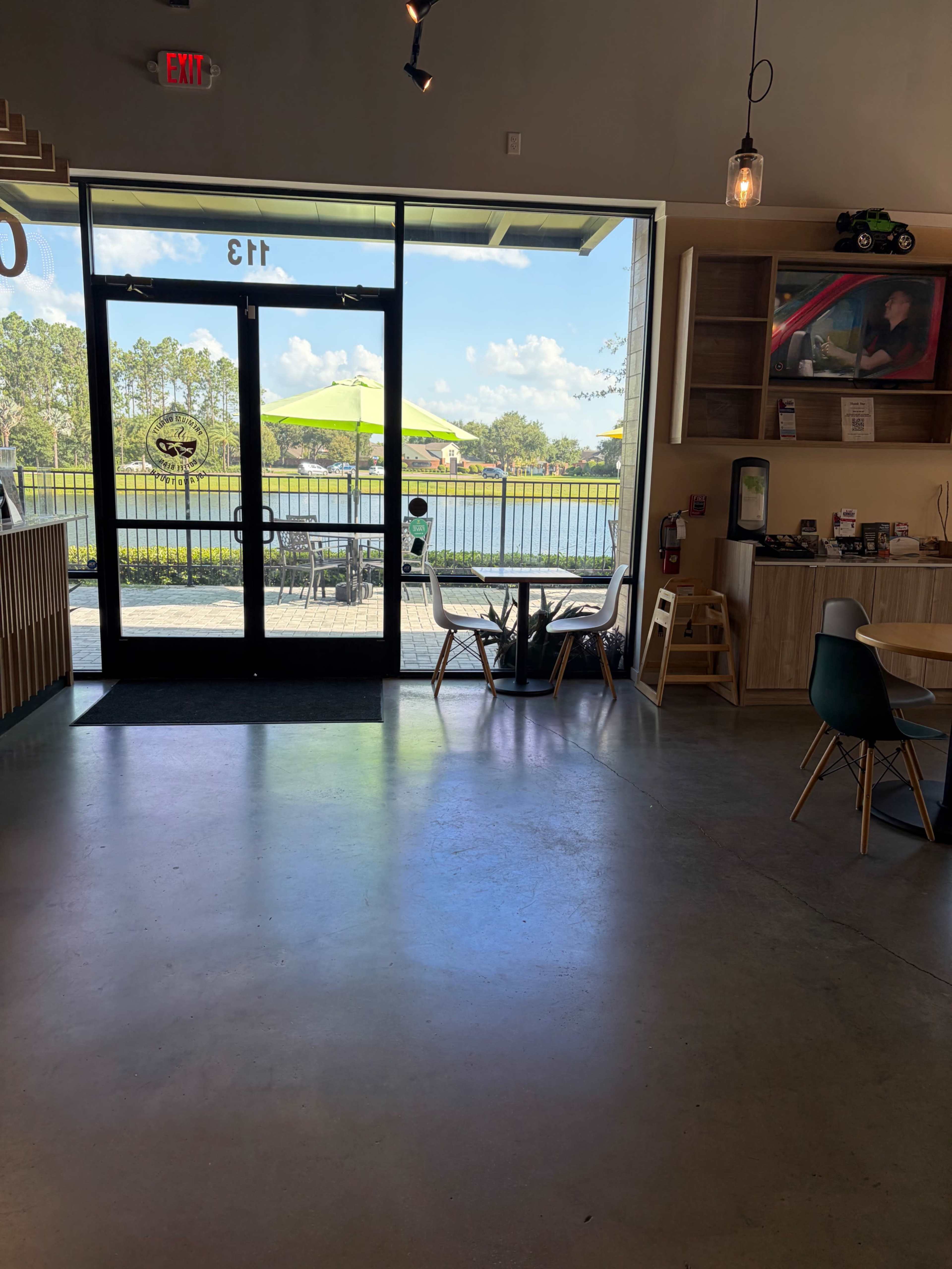 The image shows the interior of a café or restaurant with large glass doors facing a patio area, where outdoor seating and a lake are visible in the background.