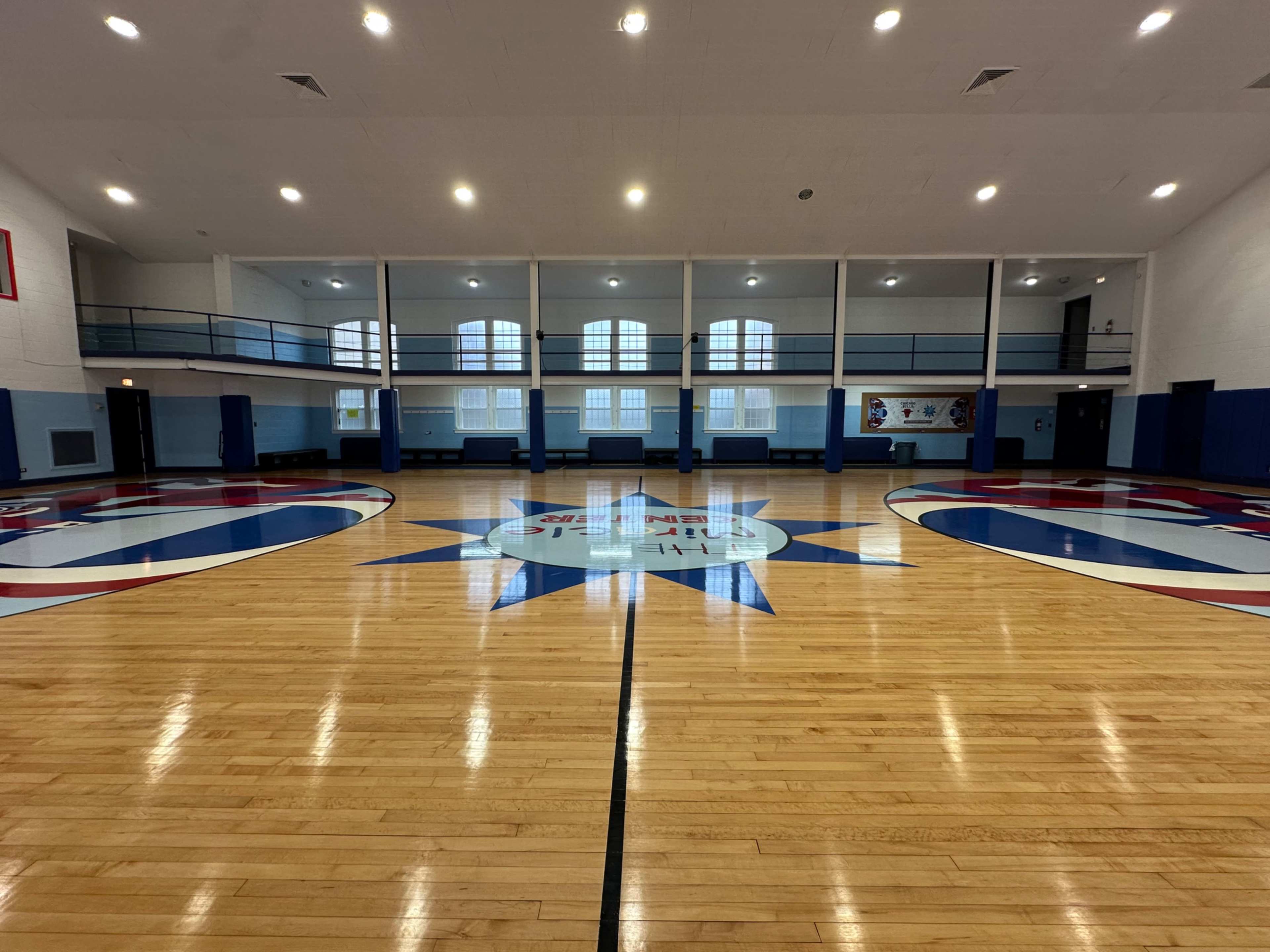 The image shows an empty gymnasium with polished wooden floors and a central star design.
