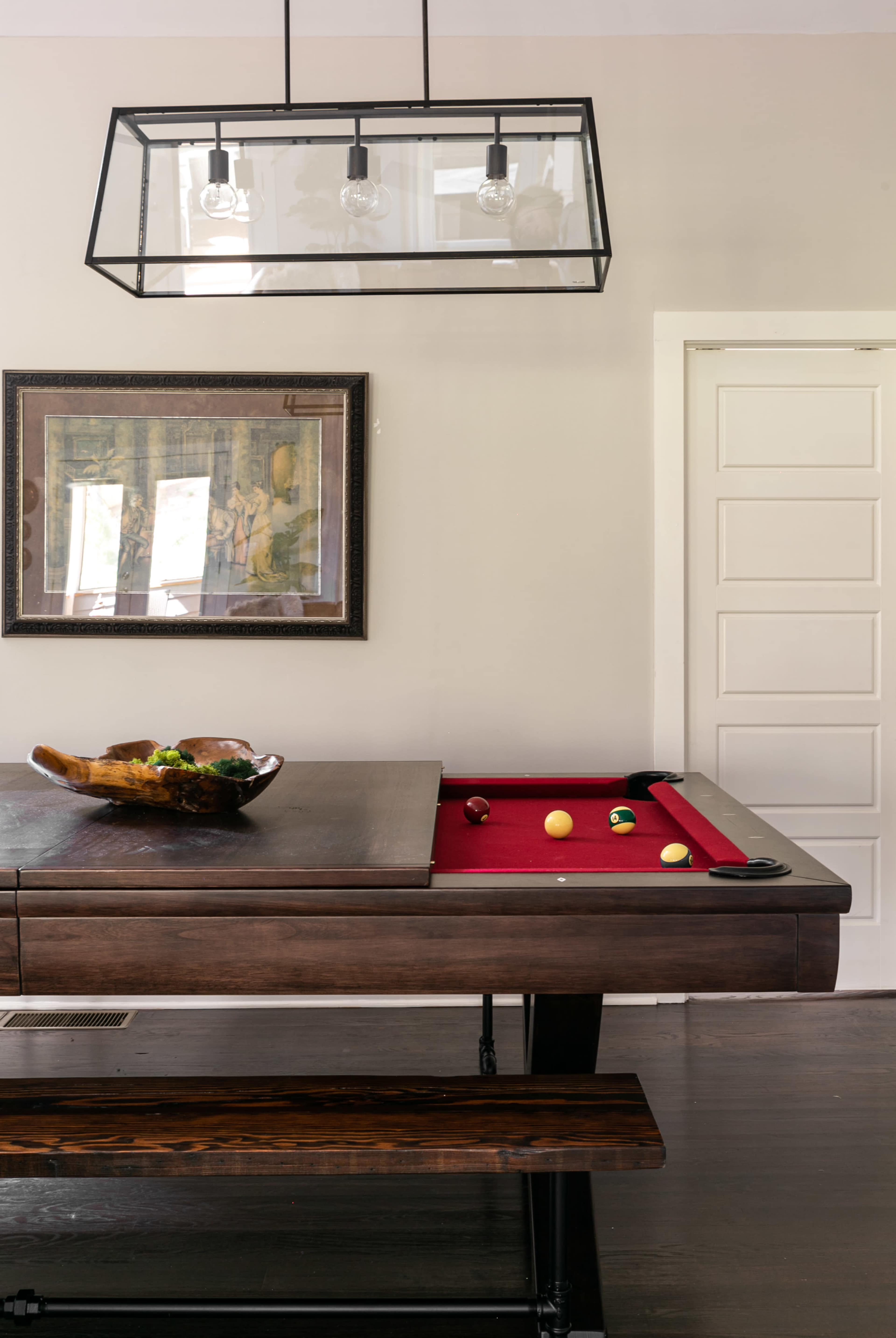 A pool table with red felt is positioned in a room featuring a framed artwork and a glass pendant light hanging above.
