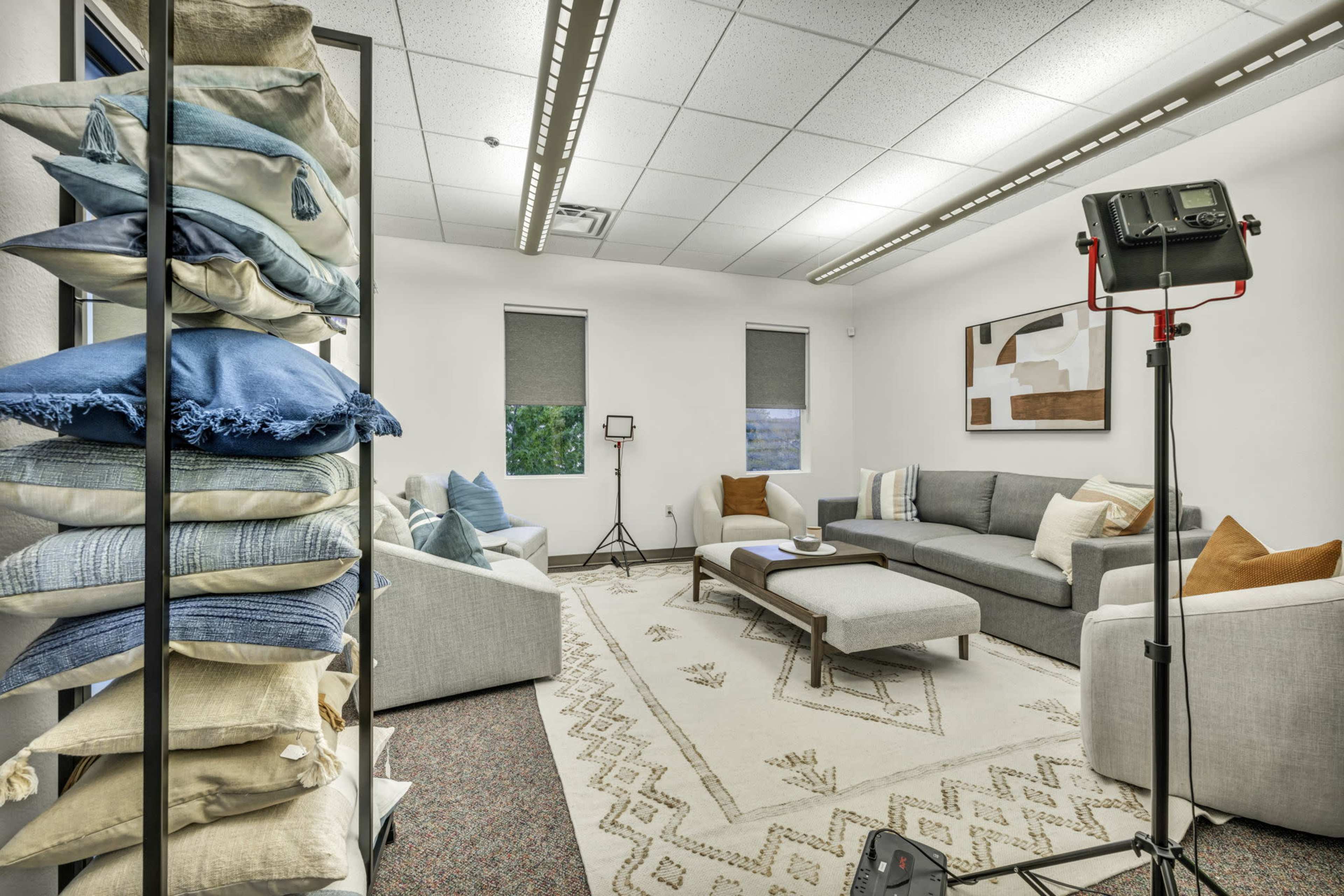 A bright room features a seating area with gray sofas, a coffee table, and a stack of colorful decorative pillows on one side.