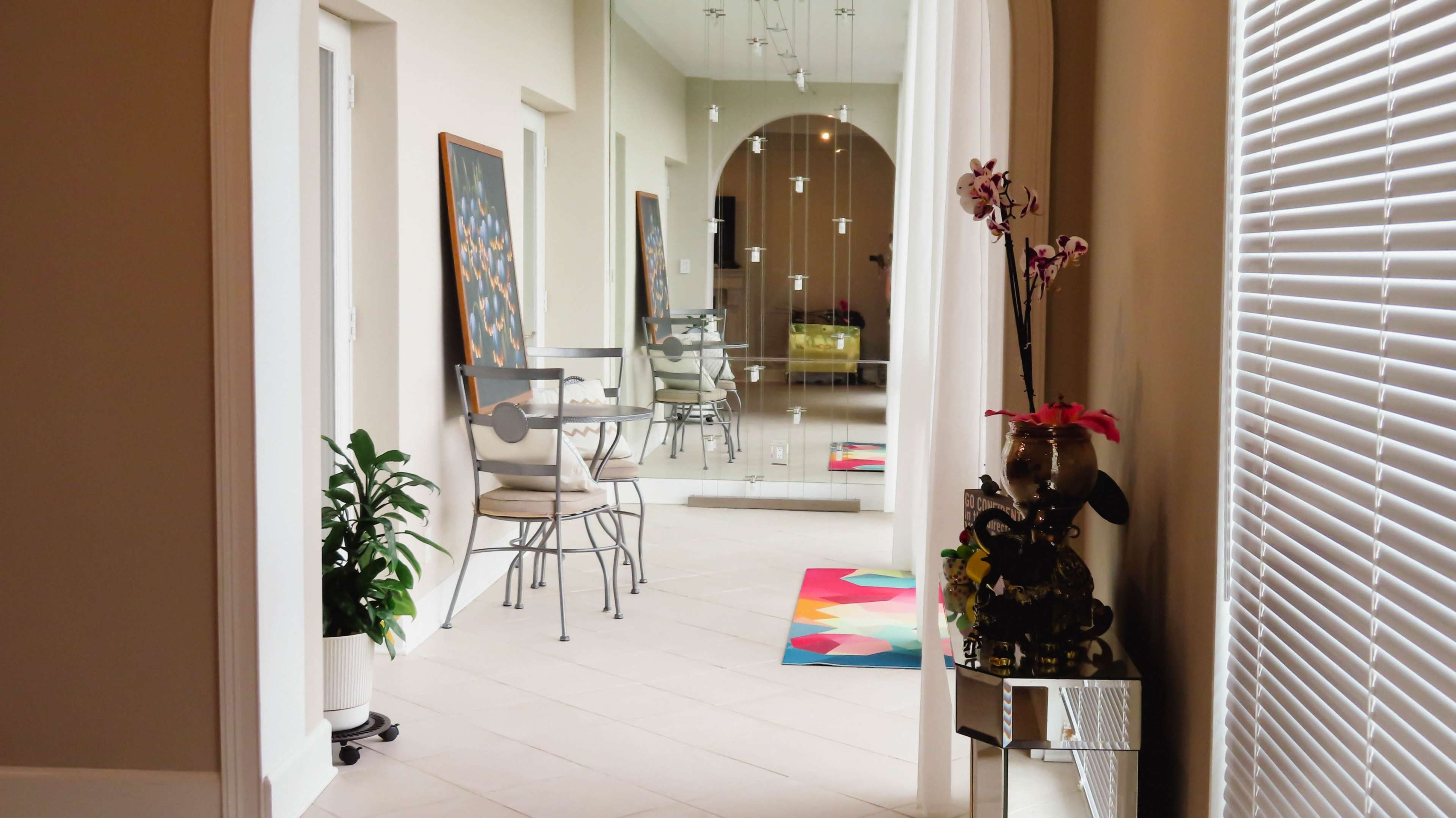 A hallway leading to a room with a mirrored wall and two chairs adjacent to a small table, decorated with plants and artwork.