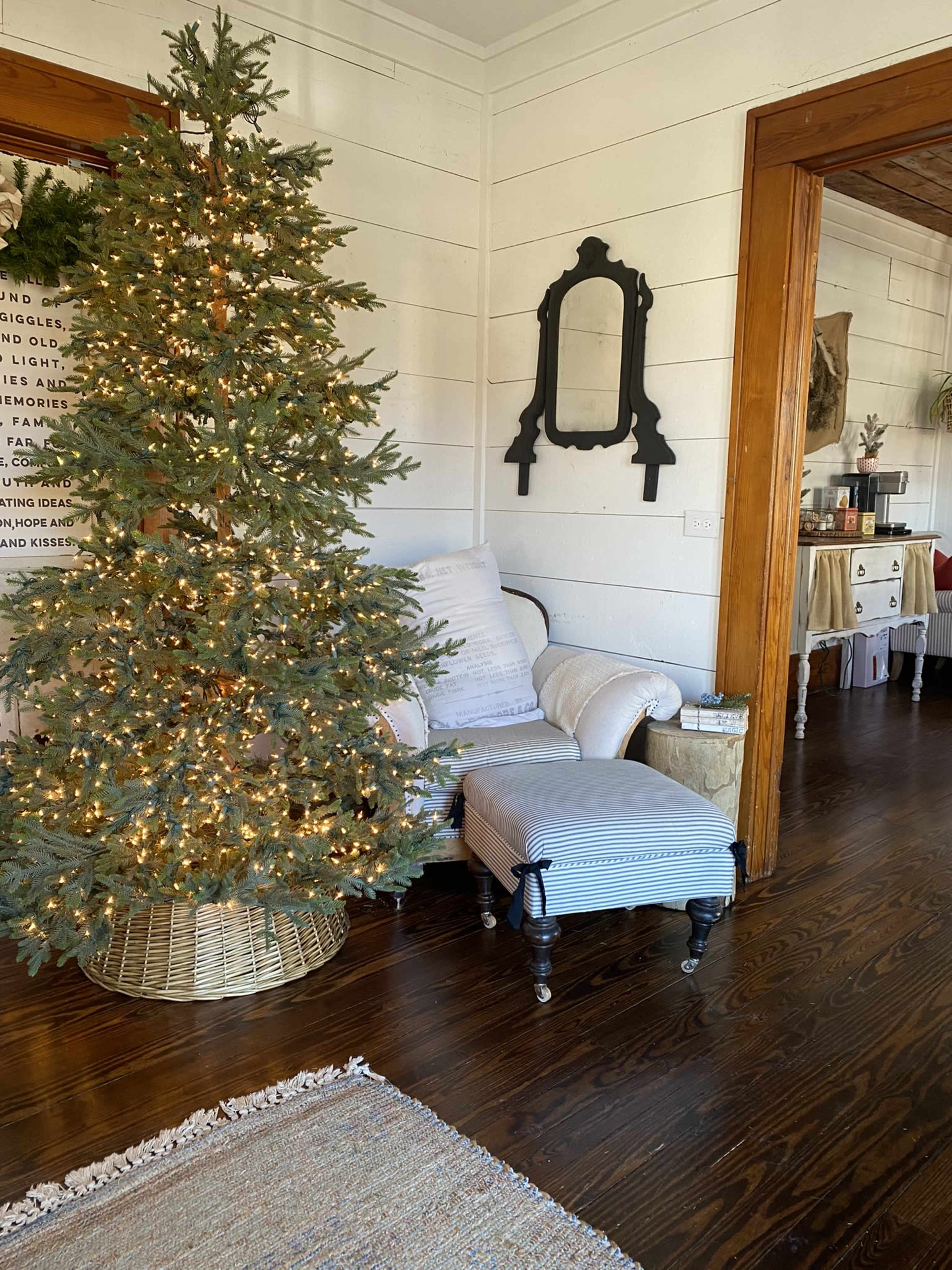 A decorated Christmas tree stands next to a chair and an ottoman in a cozy room with wooden floors and white walls.