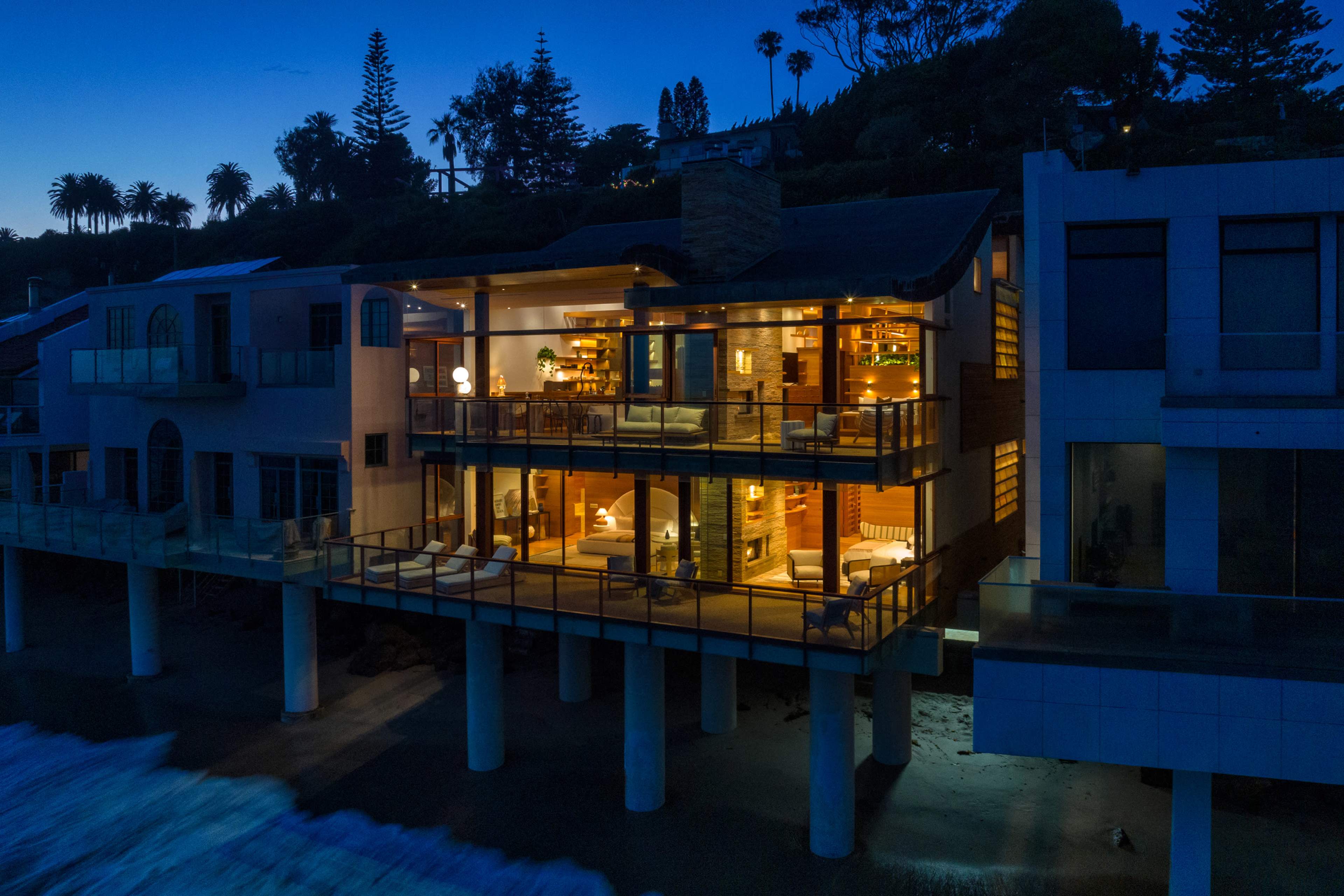 A modern house stands on stilts over the ocean, with large glass windows illuminated from within, reflecting the twilight sky.