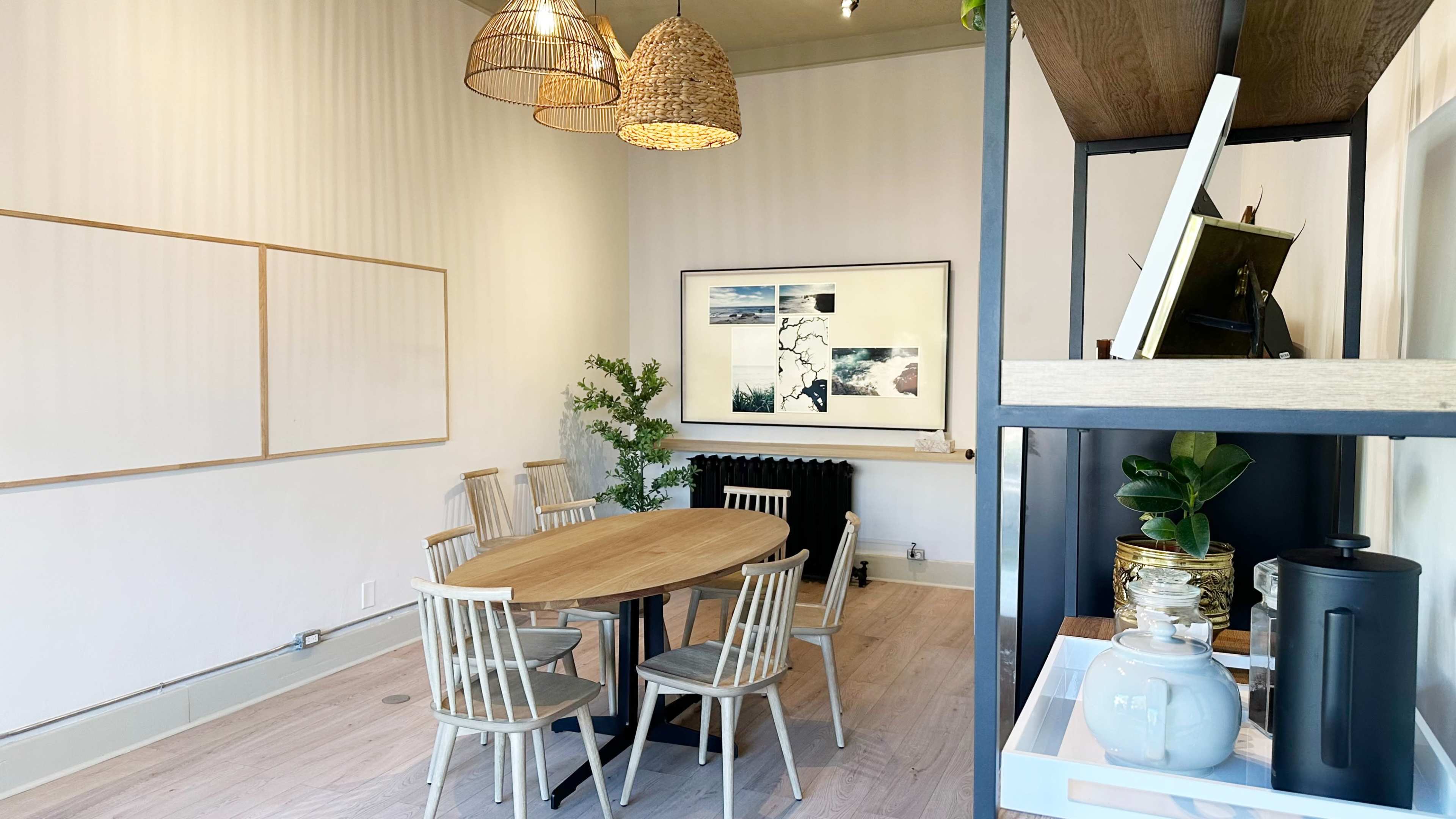 A minimalist meeting room features a round wooden table surrounded by seven chairs, with a large framed artwork on the wall and decorative plants near a radiator.