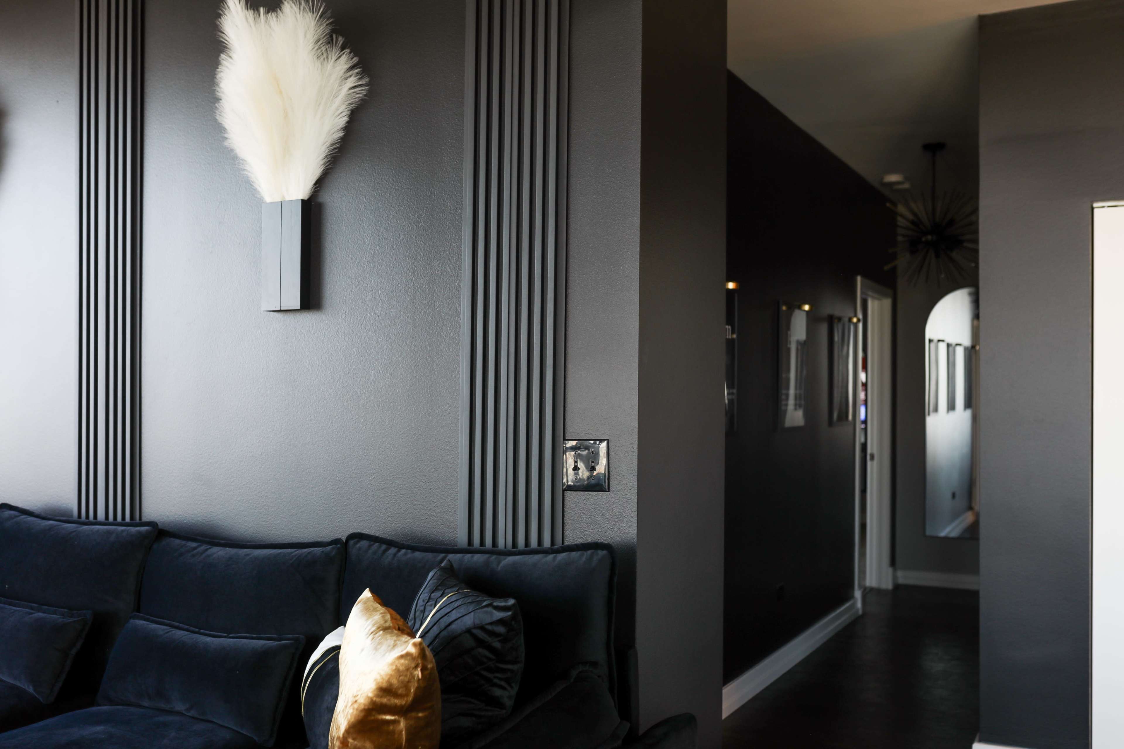 The image features a stylish living room with a dark-colored wall, a decorative wall sconce with white pampas grass, and a black couch adorned with gold and black pillows, leading to a hallway in the background.