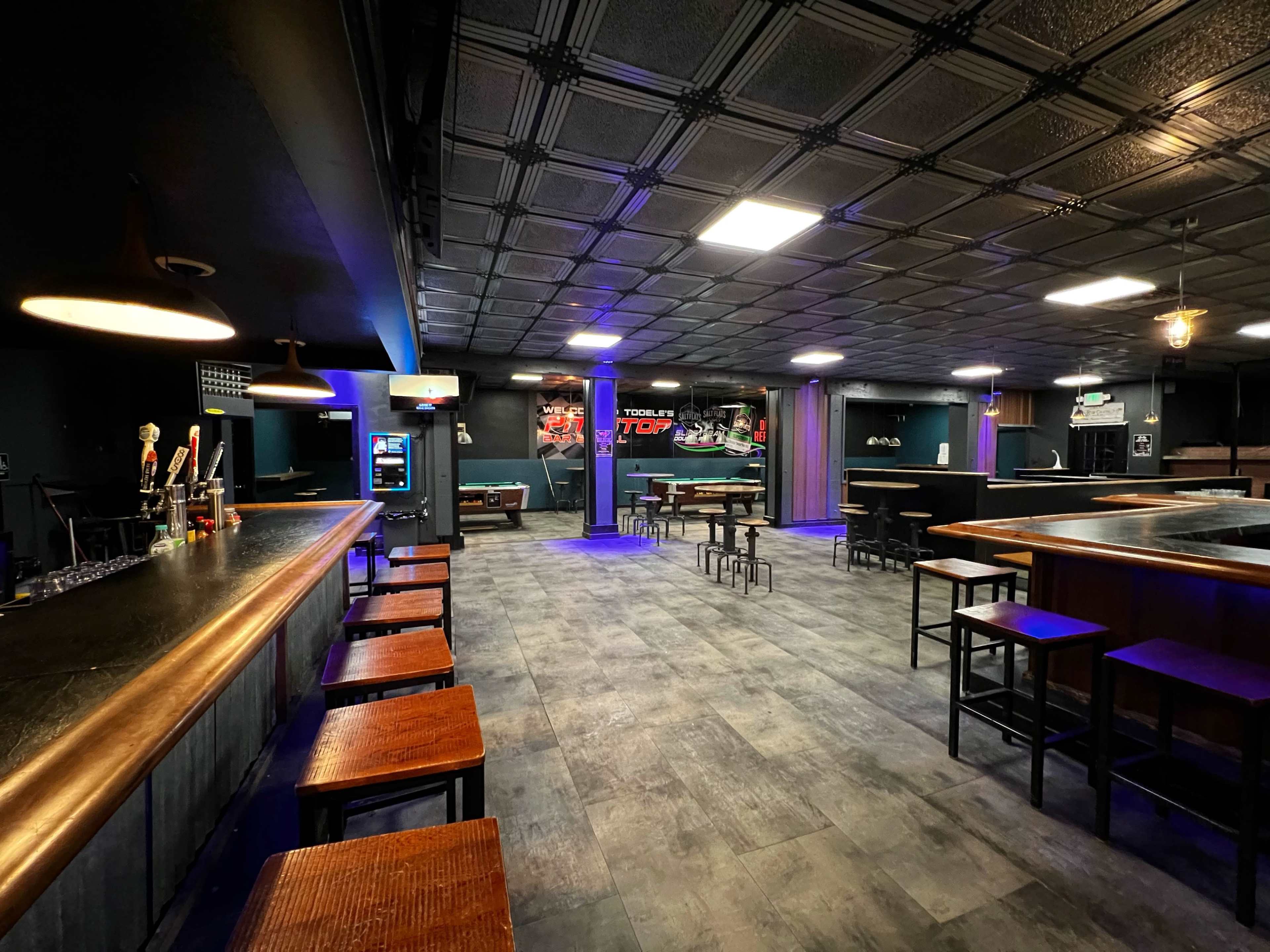 An empty bar area with a long counter, high tables, and dark walls, illuminated by blue and white lights.