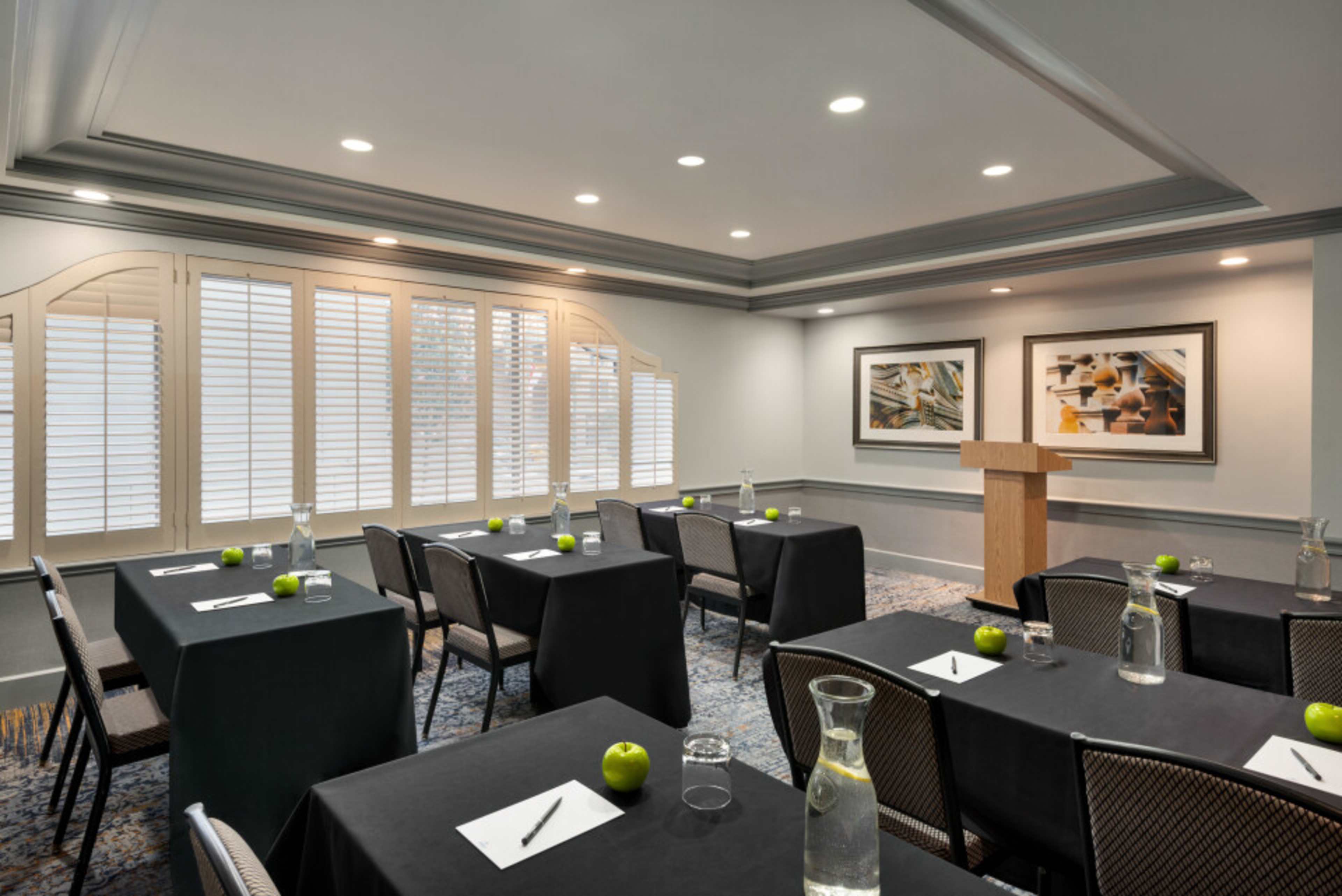 A conference room is set up with several tables covered in black cloth, each positioned with a notepad, pen, and a green apple, while a podium stands at the front near framed artwork and windows with shutters.