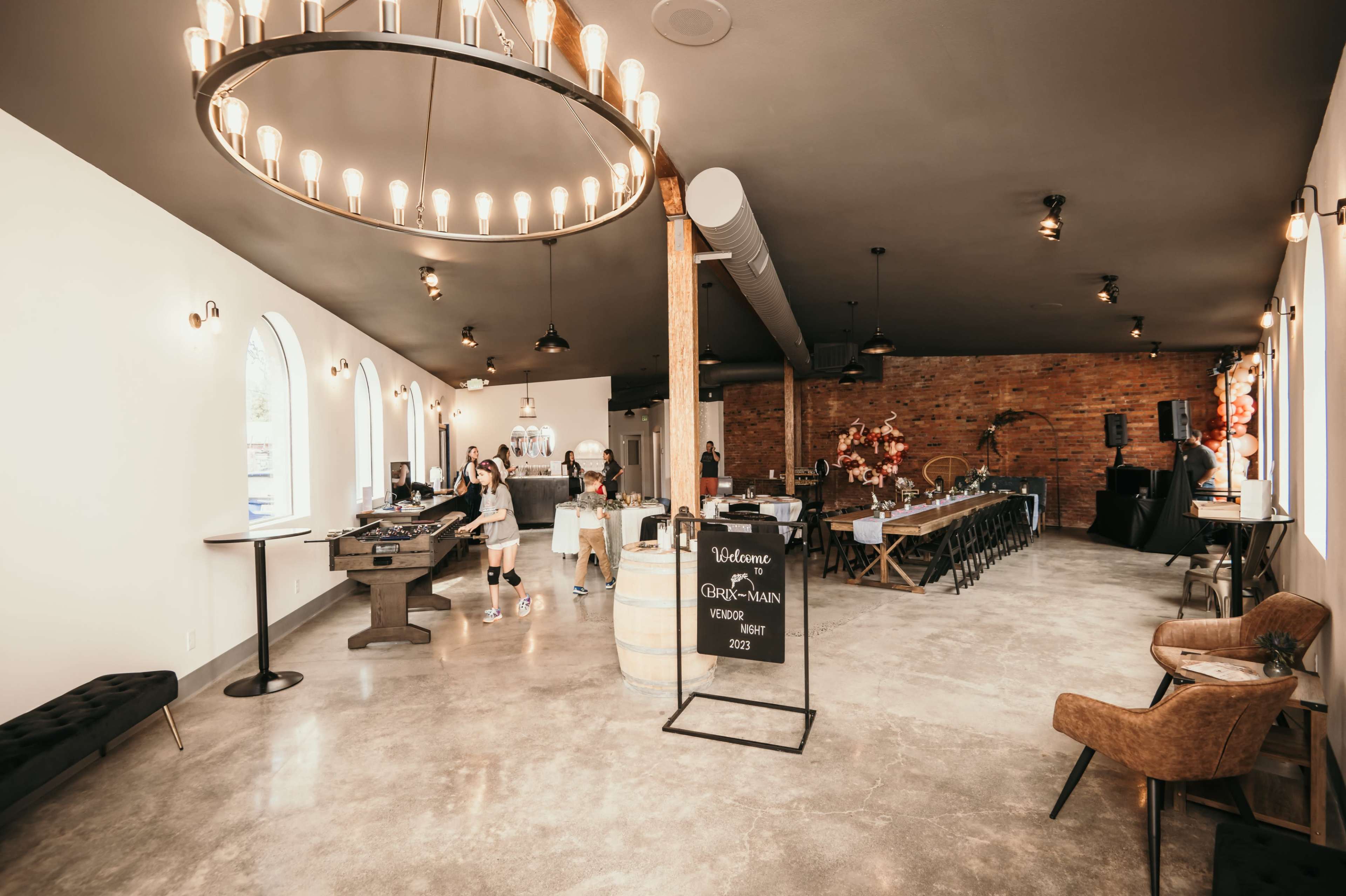 The interior of a modern restaurant features a large chandelier, wooden beams, a stone counter, and a mix of dining and seating areas.