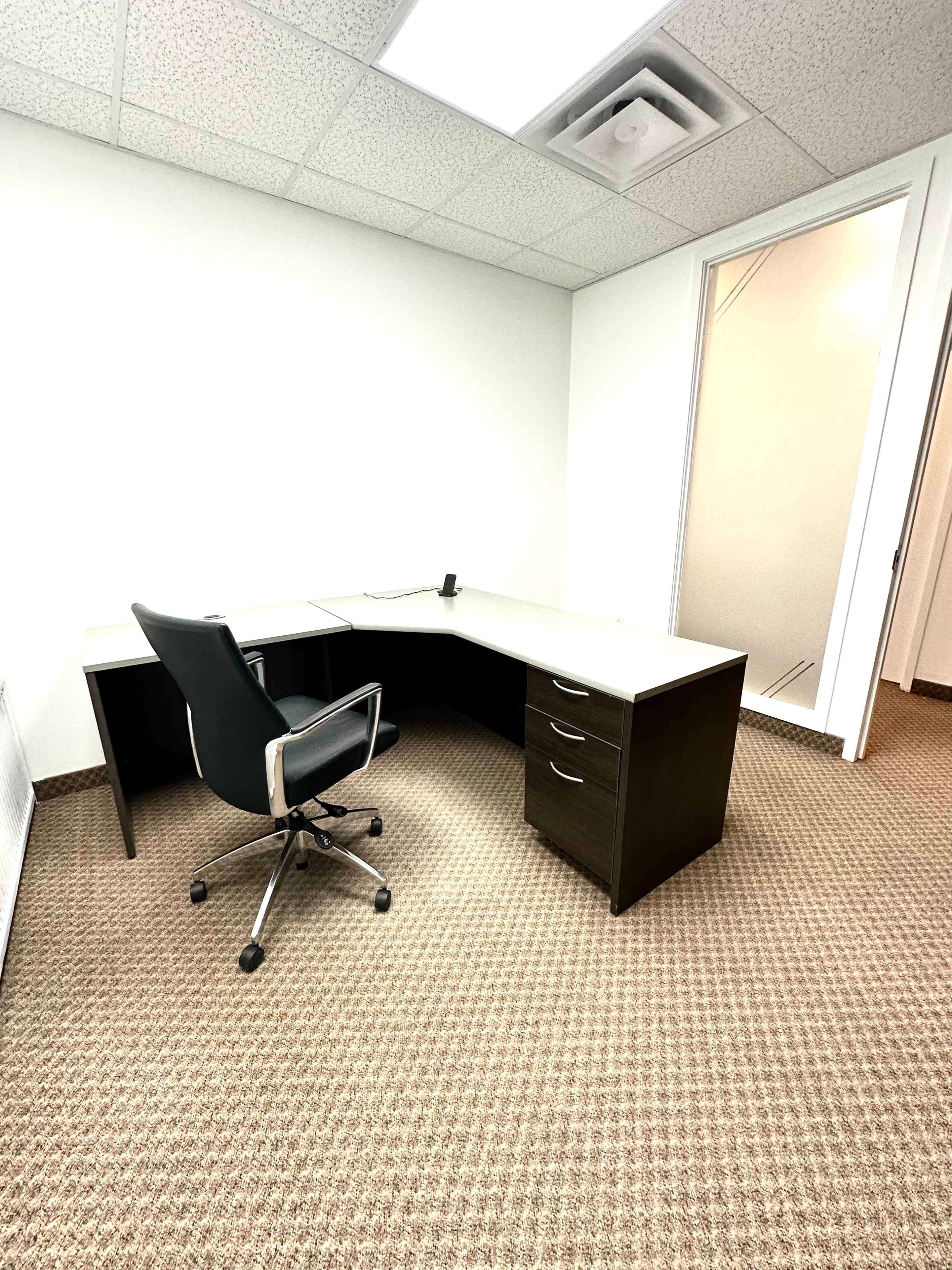 An empty office space featuring a large corner desk and an ergonomic chair on a carpeted floor.