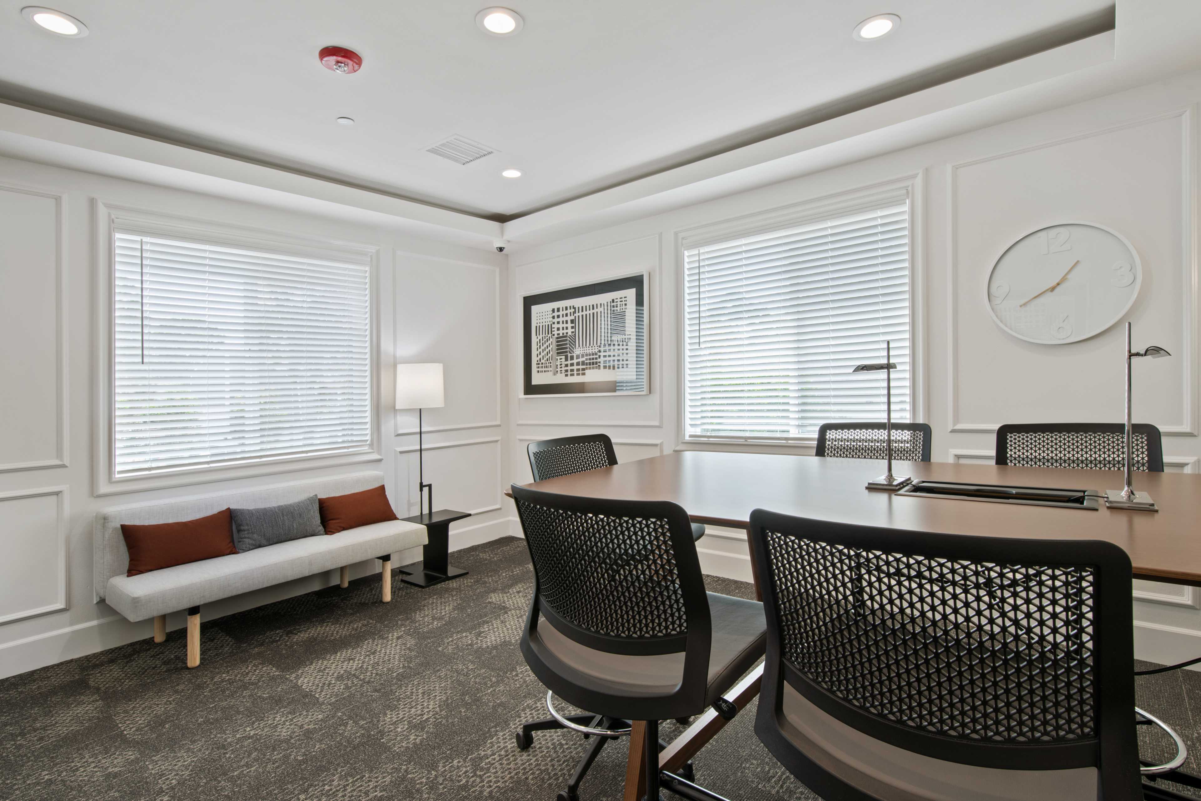 A well-lit meeting room featuring a rectangular table surrounded by black mesh chairs, a gray couch with orange cushions, and large windows with white blinds.