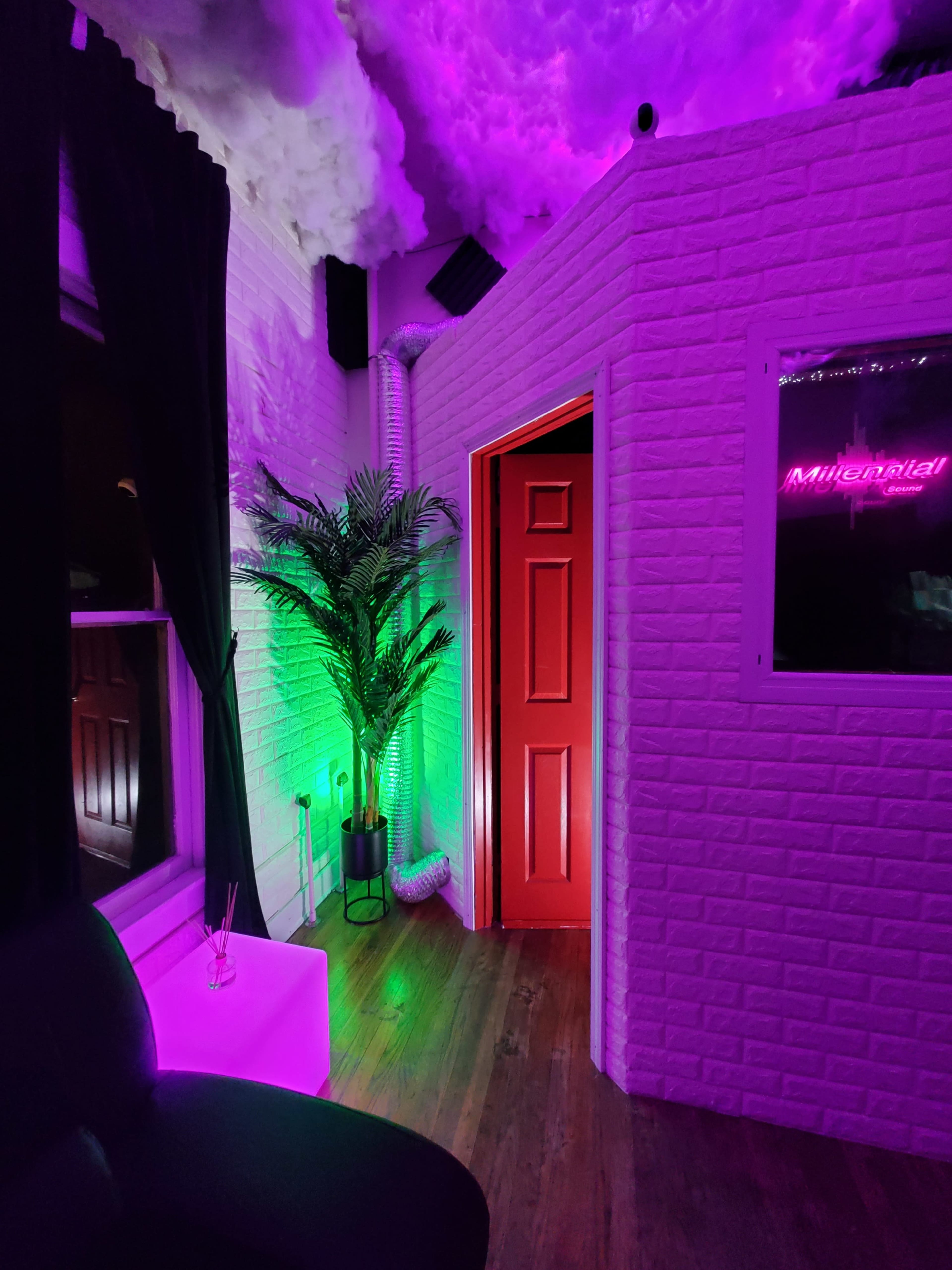 A room with a red door leading to another space, lit by purple and green lighting, with a plant and a small table in the foreground.