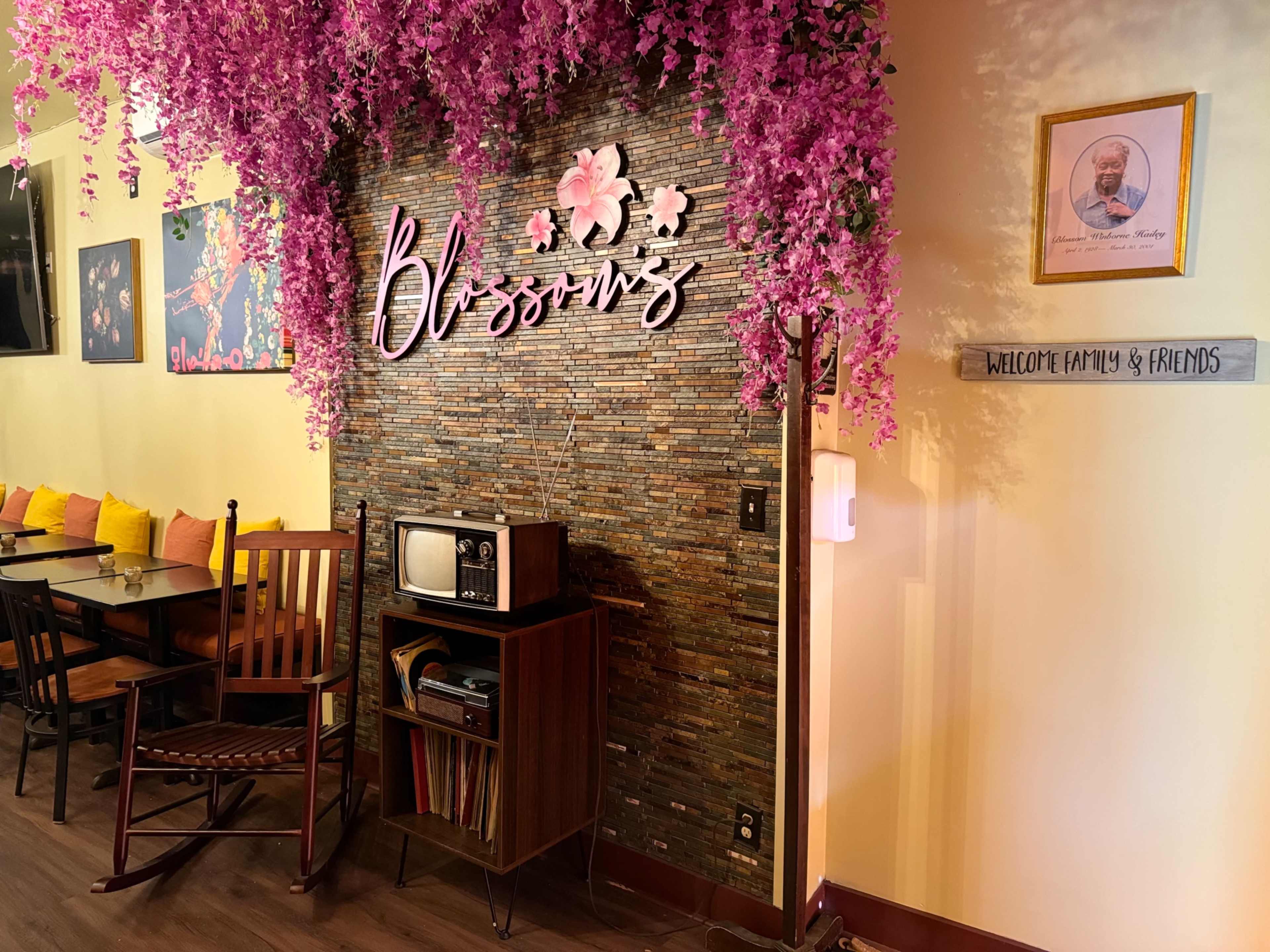 The image shows a cozy café interior with a stone wall adorned with artificial pink flowers and an old-fashioned television set beside a sign welcoming family and friends.