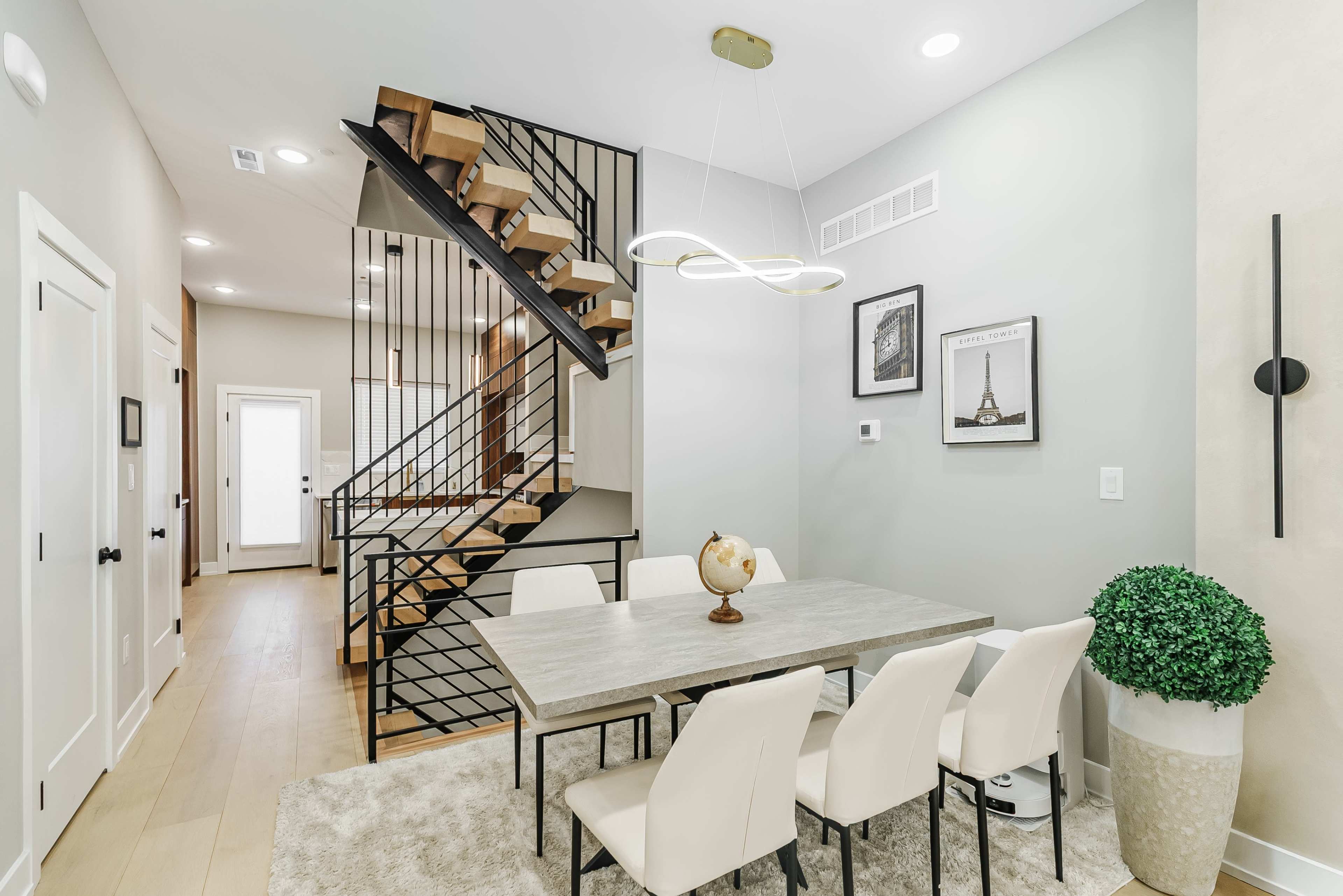 The image shows a modern dining area with a large table and white chairs, situated adjacent to a staircase with a black railing.