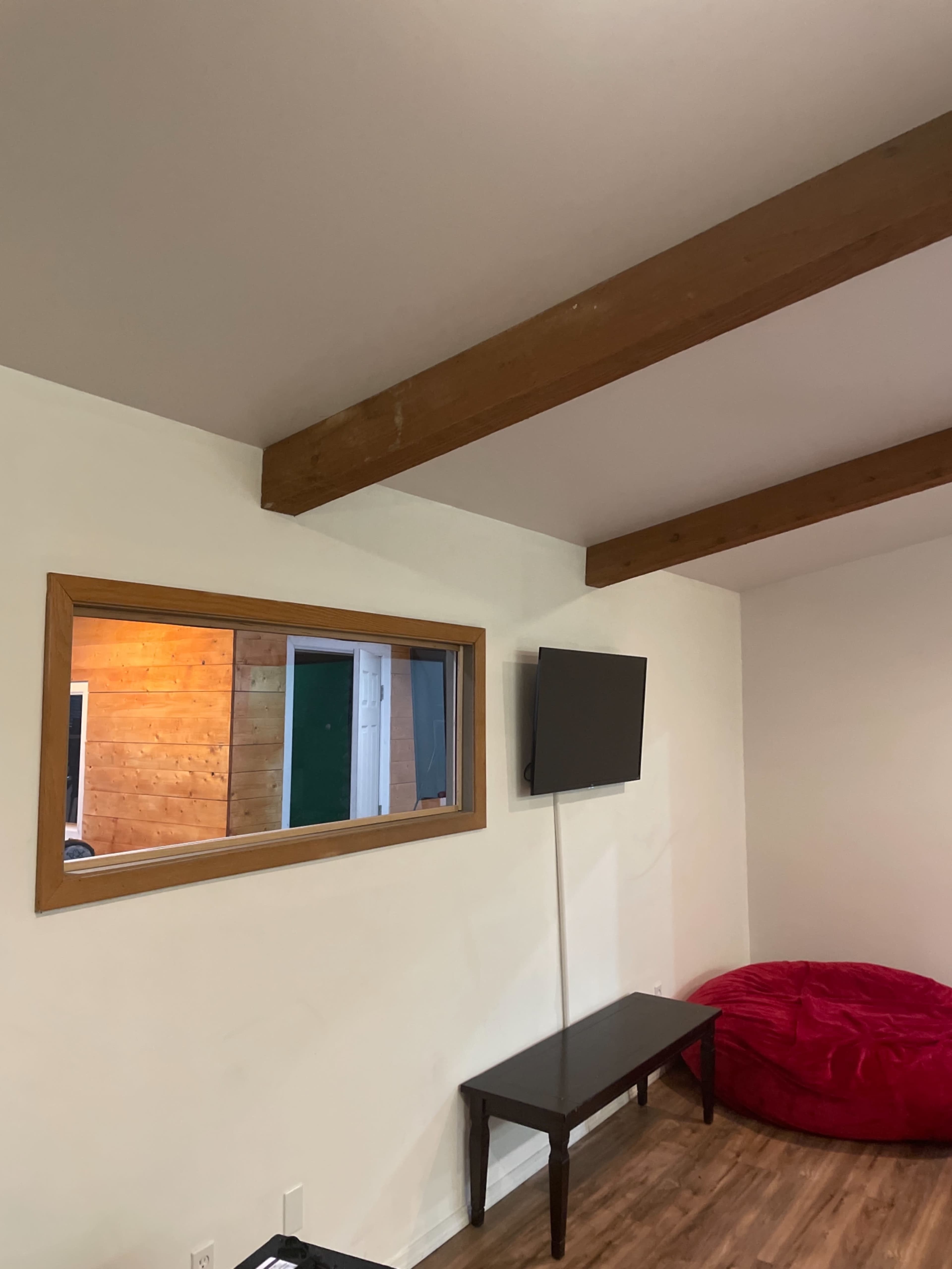 A room with a wooden beam ceiling, a wall-mounted TV, a low table, a red bean bag chair, and a window reflecting an illuminated wall.