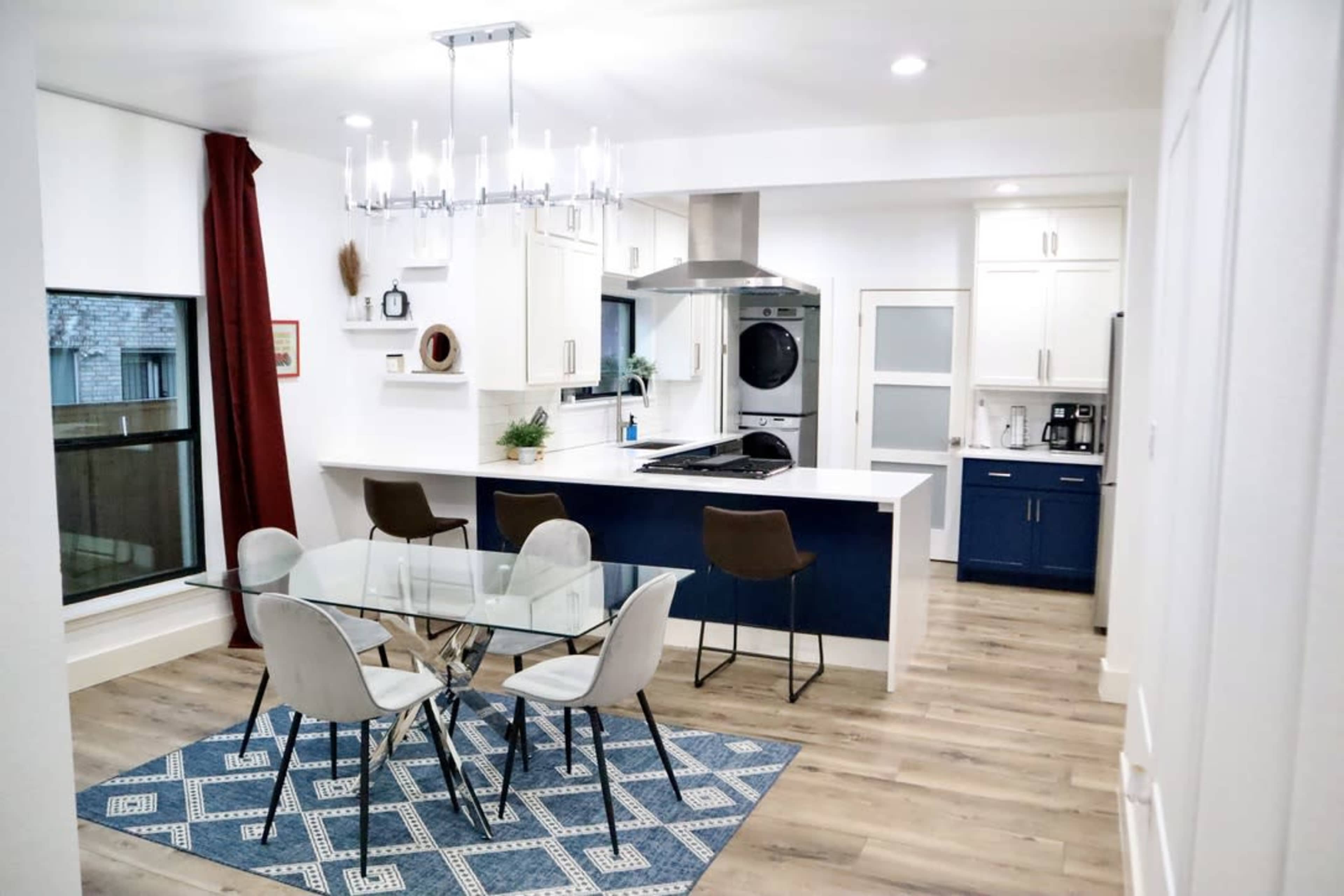 The image shows a modern kitchen and dining area with a glass dining table, four chairs, and a kitchen island equipped with a stove and overhead lighting.