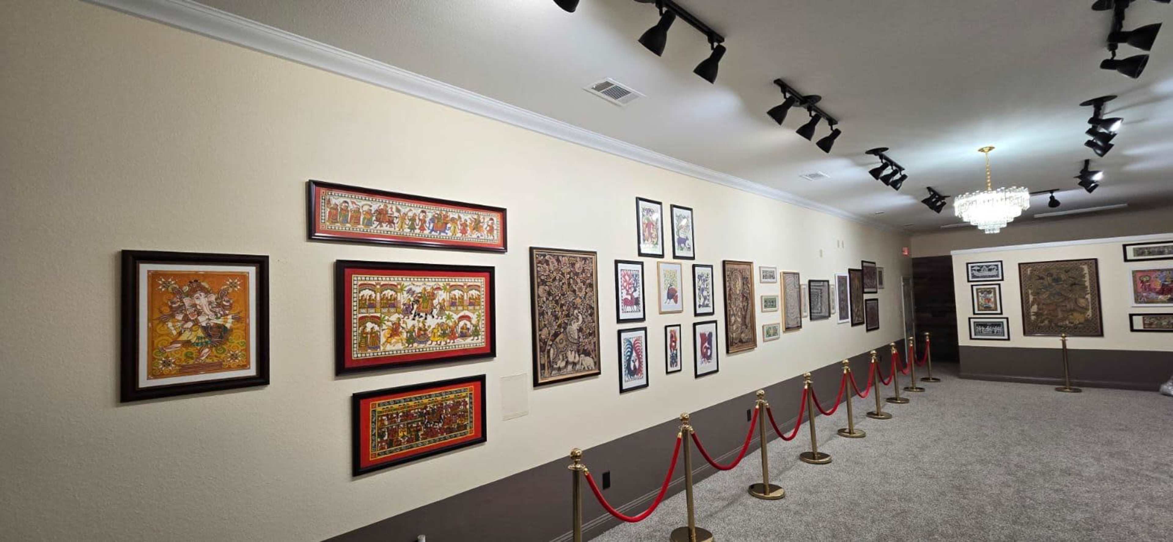 The image shows an art gallery with various framed paintings displayed along a wall, and a red rope barrier in front.