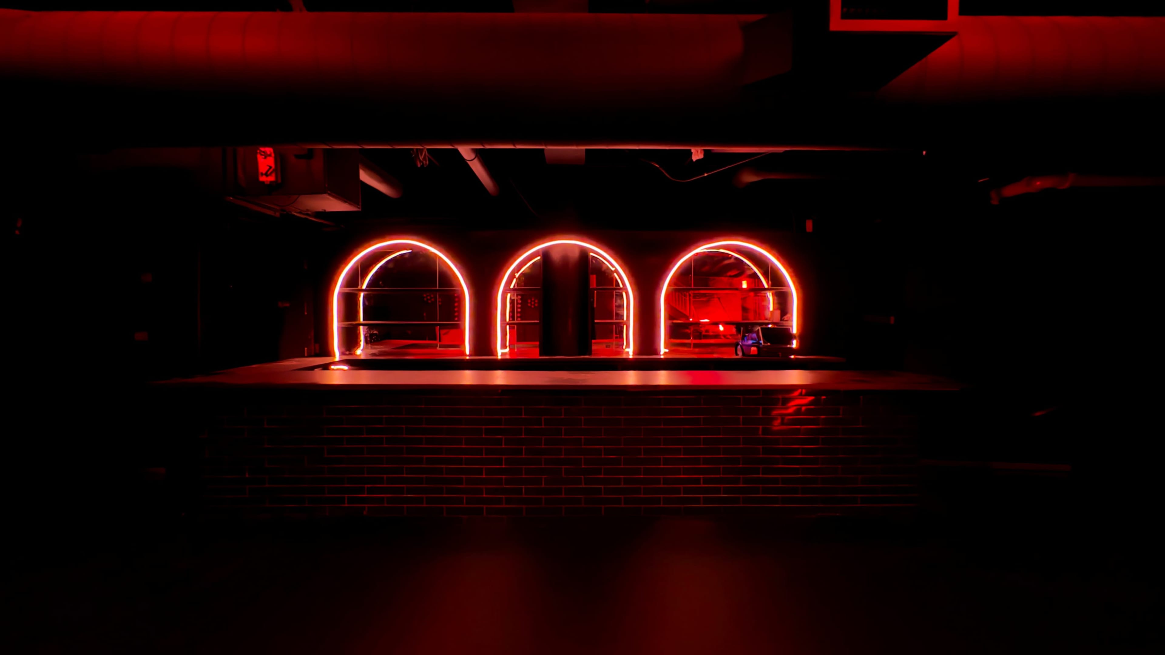 The image shows a dimly lit bar with a brick counter and three arch-shaped neon red light fixtures.